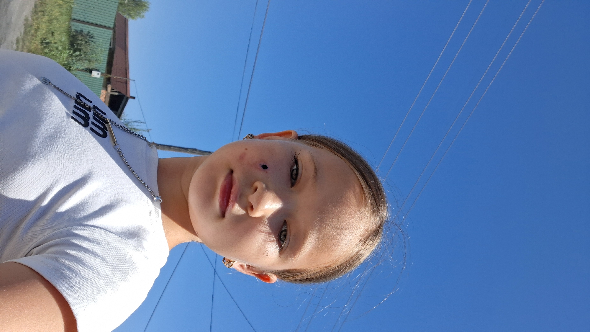 This is a selfie taken outdoors during the day, under a clear blue sky. The subject is a young girl, looking at the camera. She is wearing a white t-shirt with a silver chain around her neck. The t-shirt has black text on it, which appears to be a logo. The girl has light brown hair, and her face is illuminated by the sun. She has a beauty mark on her left cheek, and a small, possibly temporary, dark mark or drawing on her nose. She is also wearing hoop earrings. In the background, there is a green building with a brown roof, possibly a shed or outbuilding, and some trees. Power lines are visible stretching across the sky. The image is oriented sideways, suggesting it was taken with the phone held vertically. The bright sunlight and clear sky indicate fair weather. The scene appears to be in a rural or semi-rural setting, given the presence of the outbuilding and trees.