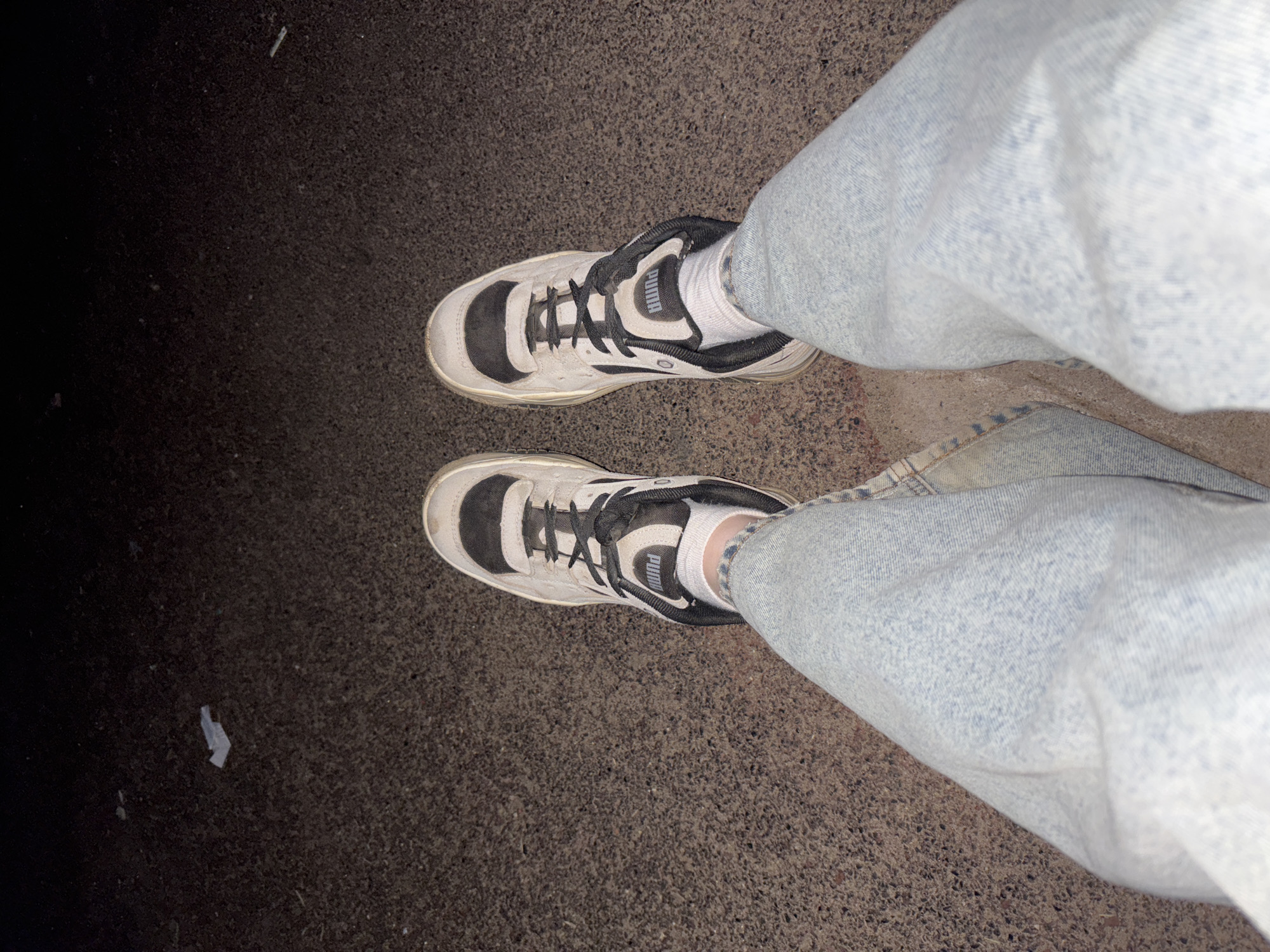 The image shows a first-person, downward-facing view of a person wearing light-wash denim jeans and black and white Puma sneakers with white socks. The person is standing on dark asphalt. The lighting is dim, suggesting an evening or night setting. A small piece of white litter is visible on the asphalt to the left of the frame. No location information is discernible, and no information is discernible to connect this image to the city of Фоки, Russia.