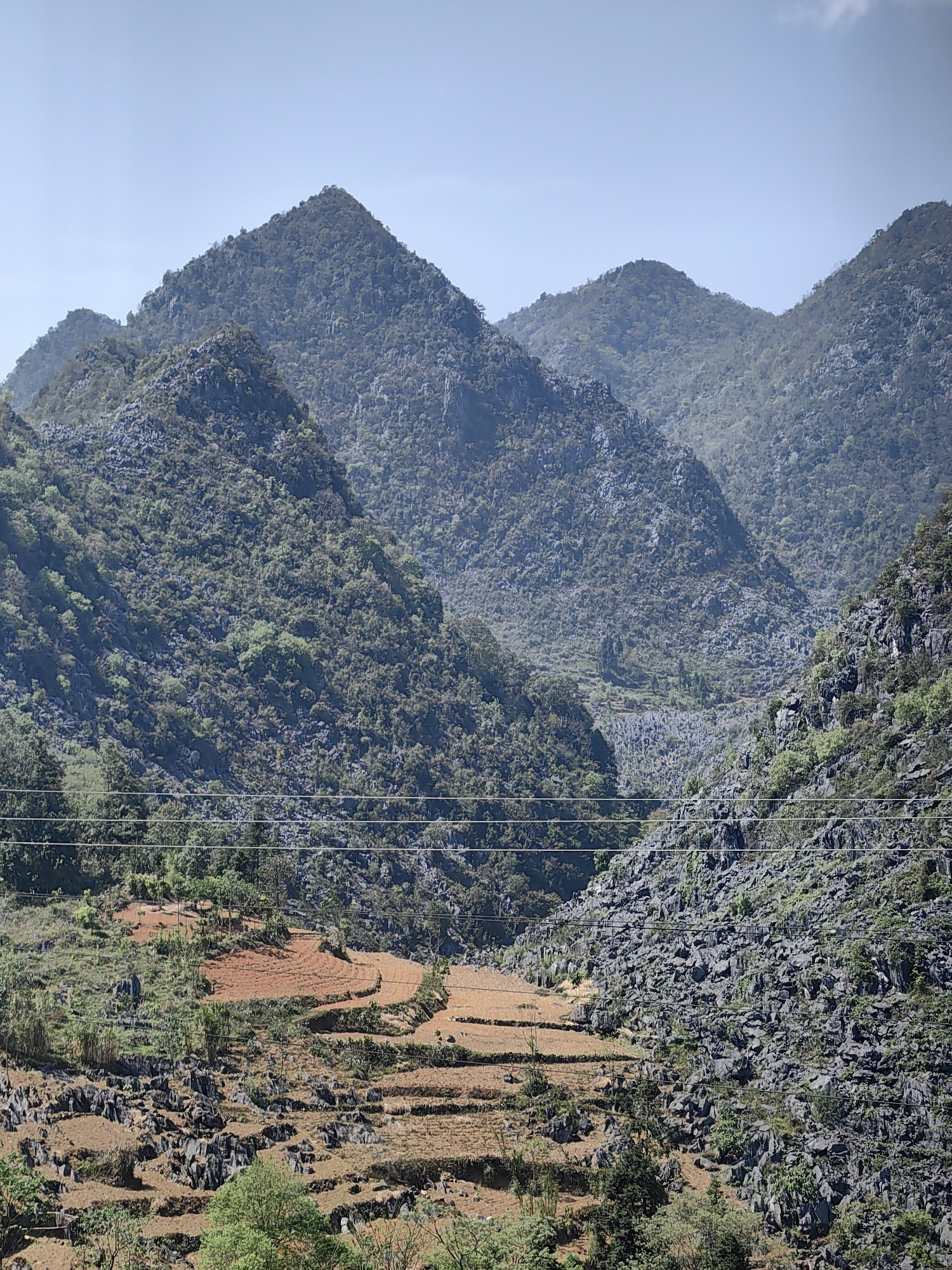 The image depicts a mountainous landscape with terraced fields in Sà Phìn Commune, Vietnam. The foreground features brown and red terraced fields interspersed with rocky outcrops and sparse vegetation. Overhead power lines cross the lower portion of the frame. The middle ground consists of steep, rocky slopes densely covered with trees and vegetation. In the background, several prominent mountain peaks rise against a clear blue sky.