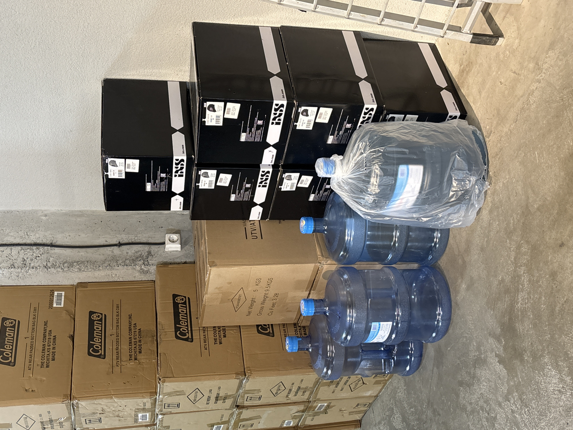 In Tbilisi, Georgia, a storage area contains stacked cardboard boxes labeled "Coleman" and several large, blue water jugs. The "Coleman" boxes appear to be new, with one visible label indicating "ATV REAR PADDED BOTTOM BAG BLK C001." Another box has a label stating "Net Weight: 5 KGS Gross Weight: 9.5 KGS Cu Feet: 3.28."
To the right of the "Coleman" boxes, a stack of black boxes with white lettering is visible. These boxes are labeled "iXS.com" and appear to be packaging for motorcycle helmets, with some labels showing product codes and descriptions like "DX691-M01-FS" and "DX691-M01-LB".
In the foreground, three large, blue, reusable water jugs are present. One jug is partially covered in clear plastic. The floor is concrete, and a portion of a metal ladder is visible on the right side of the image. The background consists of a textured wall with an electrical outlet. No people are present in the scene.