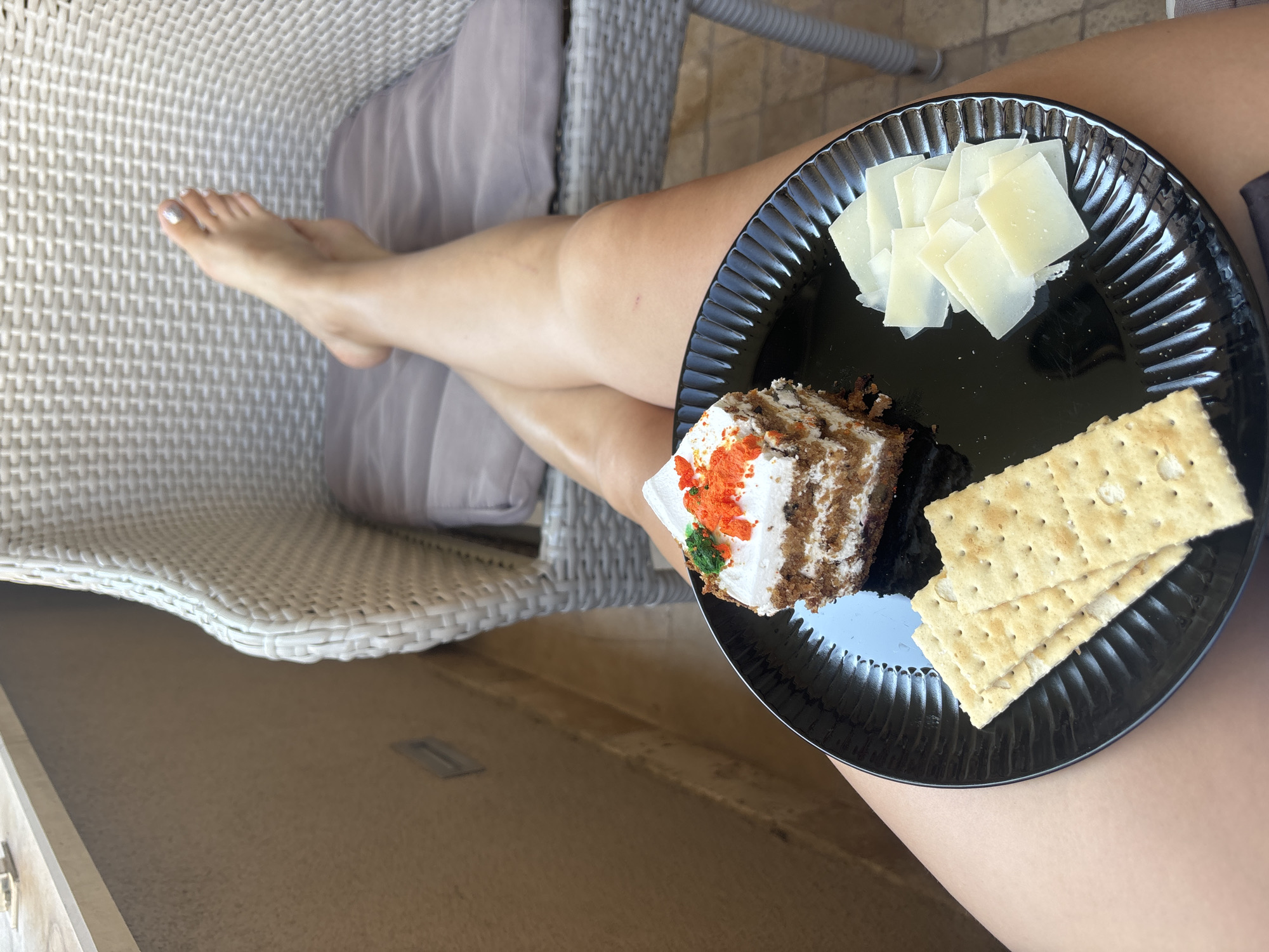 A person's legs are crossed, with one foot propped up on the seat of a rattan chair. The foot has a silver glitter polish on the toenails. In the foreground, a black plate holds a slice of carrot cake, several crackers, and slices of cheese. The scene appears to be on a balcony, with a light-colored textured wall in the background and a view of the city of Budva, Montenegro, likely from an apartment or hotel.