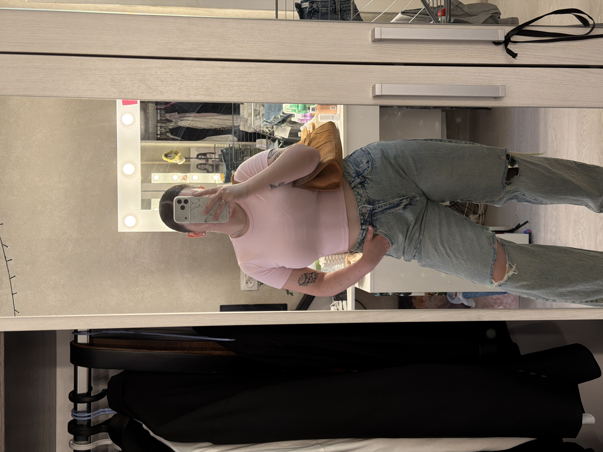 A reflection of a woman is captured in a closet mirror. She is holding a cell phone in her right hand, her arm bent at the elbow, as if taking a selfie. The woman is wearing a pink t-shirt and light blue ripped jeans. The tan purse is slung over her shoulder. The closet has multiple shelves with folded clothing, shoes, and accessories. A dressing table with vanity lighting is visible in the background. The photograph appears to have been taken indoors. Location is Chaikovsky, Russia.