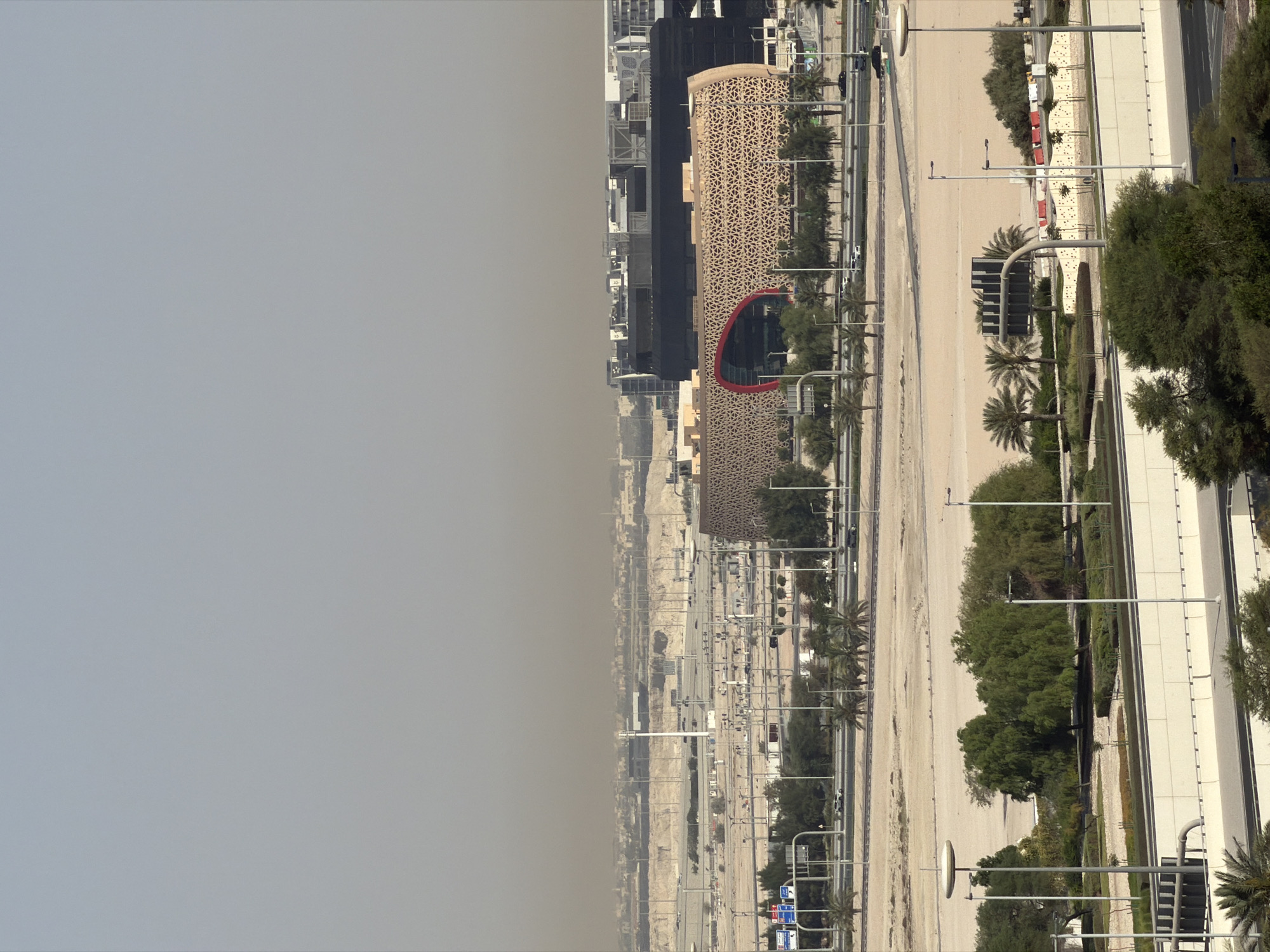 This image captures a wide view of a modern cityscape under a hazy sky, likely due to dust or pollution. The foreground features a multi-lane highway with concrete barriers and streetlights. Beyond the highway, a sandy, sparsely vegetated area is visible, dotted with palm trees and other greenery. In the mid-ground, a distinctive, curved building with a patterned facade and a large red-outlined opening stands out. Behind this building, a large, dark-colored structure with a flat roof and several smaller buildings are situated. The background reveals a vast expanse of urban development stretching towards the horizon, shrouded in atmospheric haze. Various vehicles are present on the roads, and utility poles are scattered throughout the landscape. The overall scene suggests a contemporary, possibly arid, urban environment.