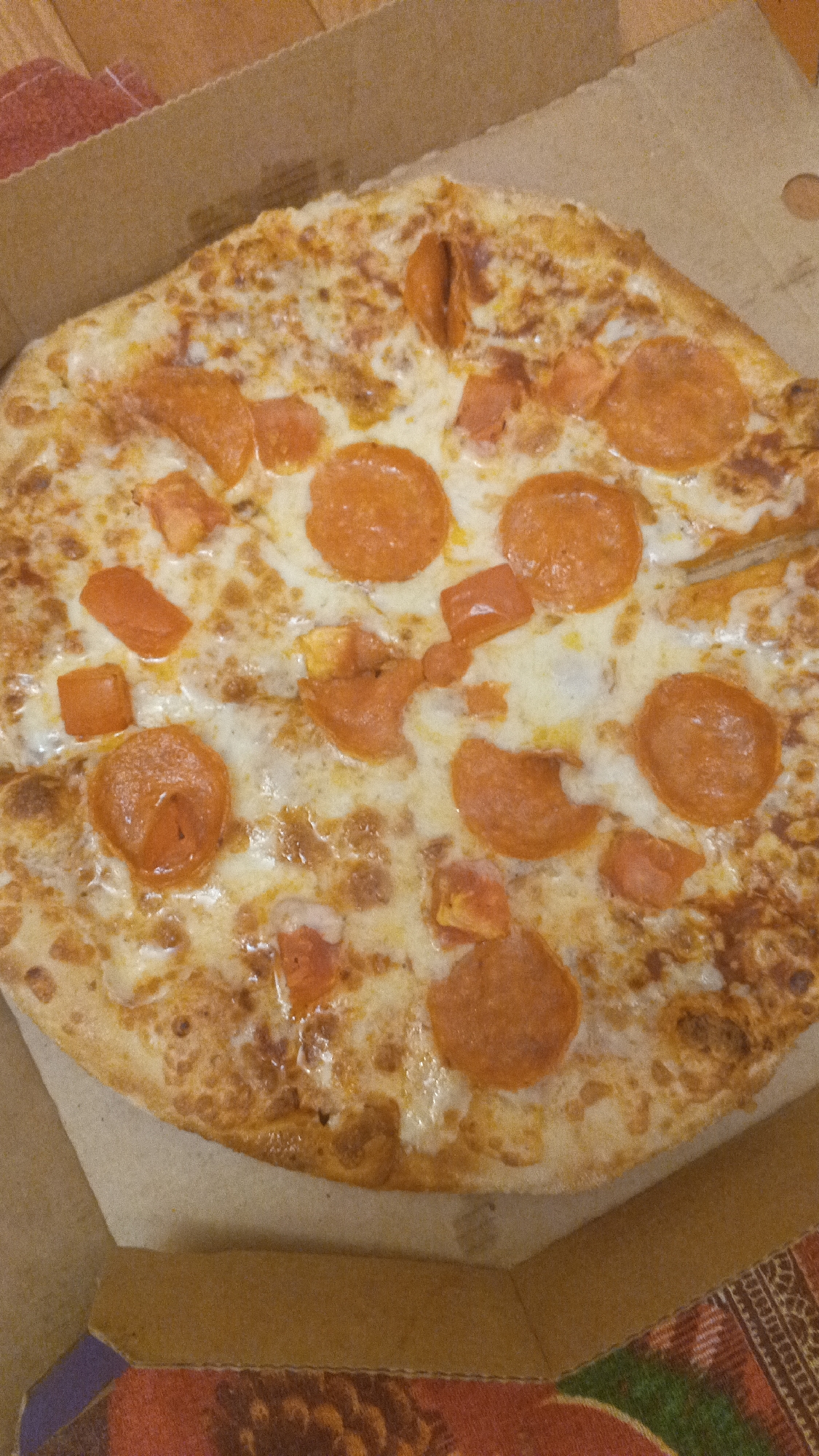 A close-up, overhead shot shows a round pepperoni pizza with cheese and tomato pieces, resting in a cardboard pizza box. The pizza is golden brown with some charring on the crust and melted cheese. Slices of pepperoni are scattered across the pizza, along with small, diced tomato pieces. The pizza box is made of corrugated cardboard and is open, revealing the pizza inside. The background shows a wooden surface and a glimpse of a colorful patterned cloth beneath the pizza box. There are no people or animals present in the image. The location in the city of Mytishchi, Russia, is not evident from the image itself.