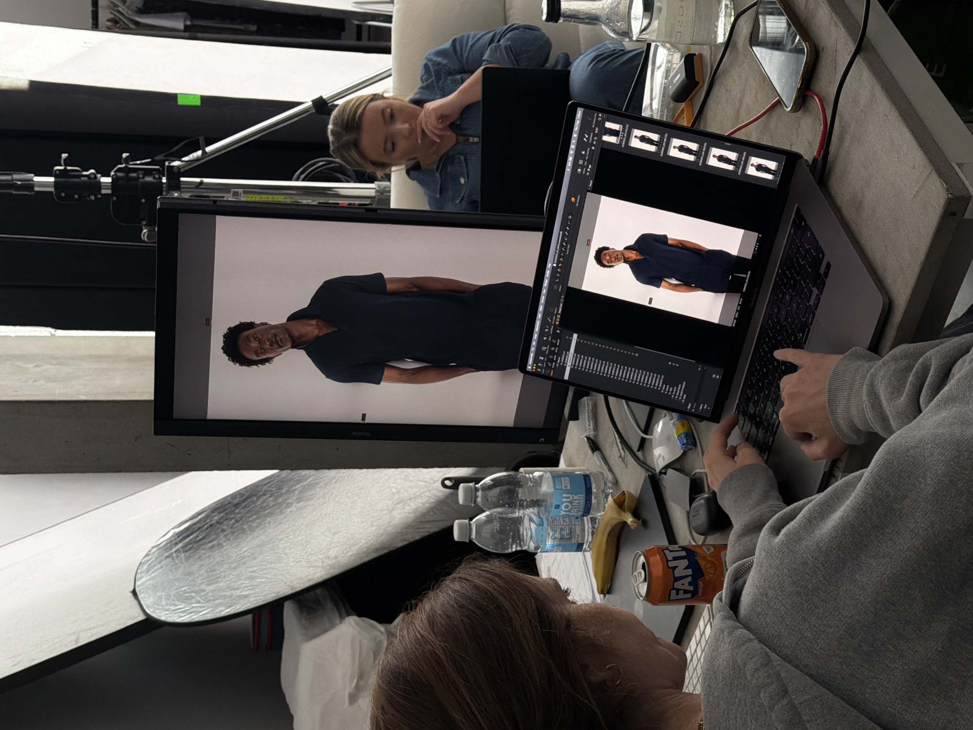 The image captures a scene likely from a photoshoot or post-production environment. Two women are present, one reclining on a white couch wearing a denim shirt, looking at a laptop on her lap. The other is seated at a concrete table or surface, operating a laptop. The laptop is connected to a large monitor displaying an image of a Black male model in a dark blue shirt and pants. The laptop screen also displays image editing software with multiple thumbnail images of the model. Other objects on the table include two bottles of water, a banana, a can of Fanta soda, a smartphone, and various cables. A photography reflector is partially visible in the lower-left corner. Behind the monitor, photography lighting equipment is visible. Based on the style of the setting and the presence of photography equipment, it's plausible that this scene is taking place in a photography studio. The image does not provide any information to confirm the location to be in the city of London, United Kingdom.