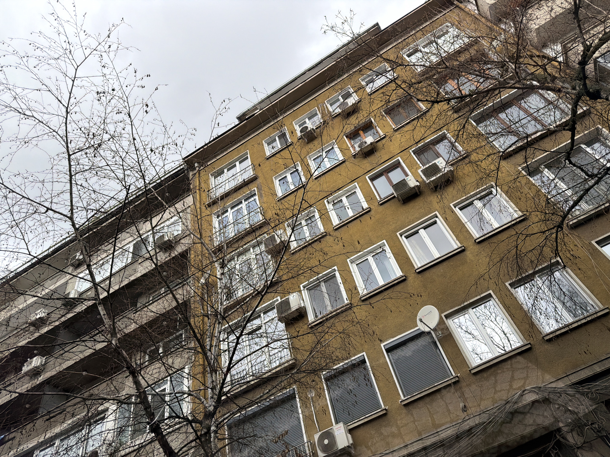 A low-angle shot depicts two buildings in Sofia, Bulgaria, set against a cloudy sky. The building on the right has a yellow facade and numerous windows, many fitted with air conditioning units. A satellite dish is visible on one of the lower floors. Trees with bare branches partially obscure the building. The building on the left is gray and features balconies.