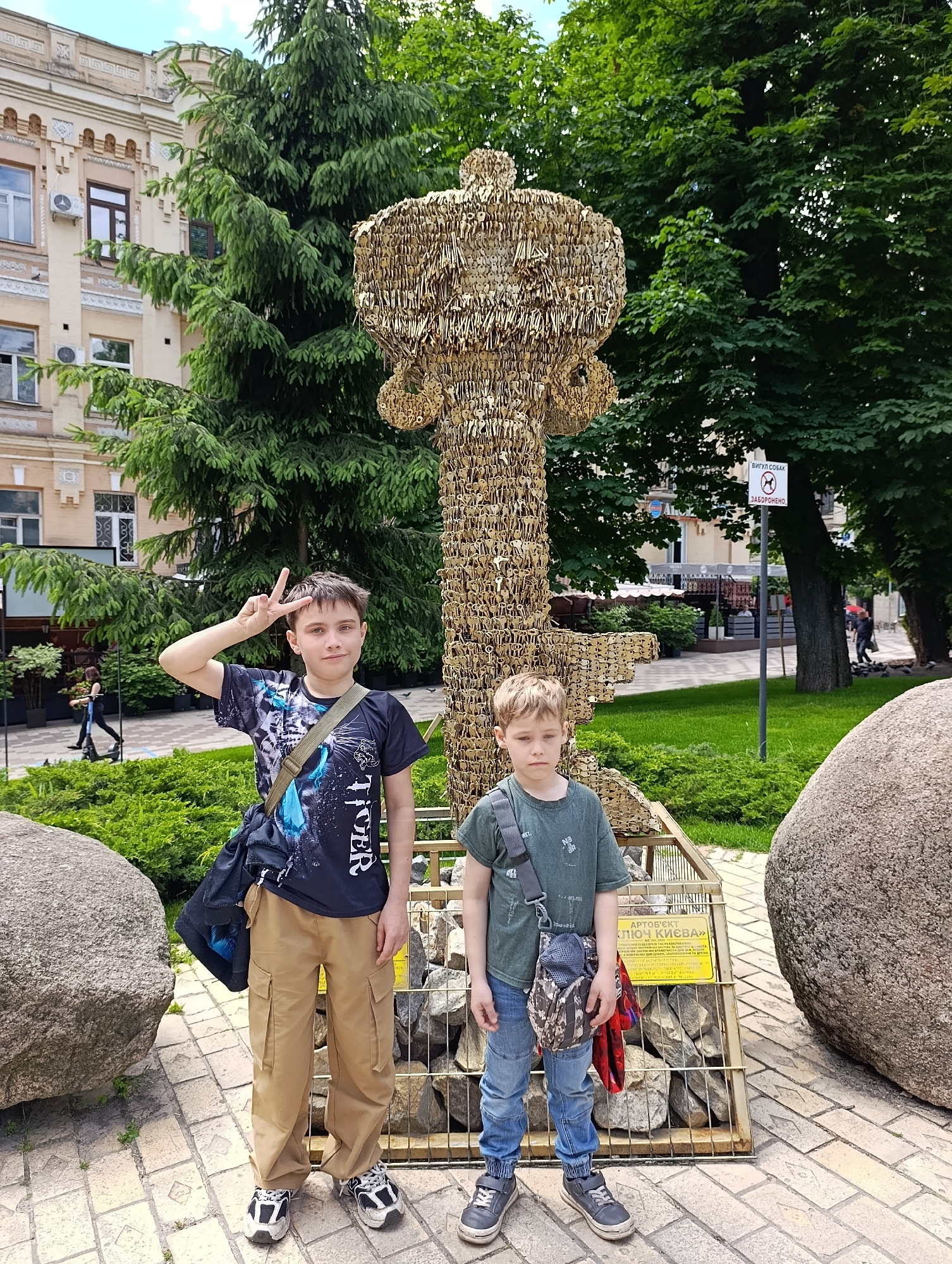 Two young boys are posing for a photo in front of a large sculpture of a key made of many smaller keys. The sculpture is situated in a park-like area with trees and grass. In the background, a building with classical architecture is visible. The younger boy is standing on the right, wearing a green t-shirt and jeans, with a camouflage-patterned bag slung across his chest. The older boy is on the left, wearing a black t-shirt with a graphic print and khaki cargo pants, with a green strap bag over his shoulder and making a peace sign with his fingers. A sign with Cyrillic lettering is visible behind the younger boy. The ground is paved with bricks, and large rocks are placed around the base of the sculpture. The overall setting suggests an outdoor tourist attraction in a city. This key sculpture is part of the "Key of Kyiv" art object, located on Konshenka Street in Kyiv, Ukraine.