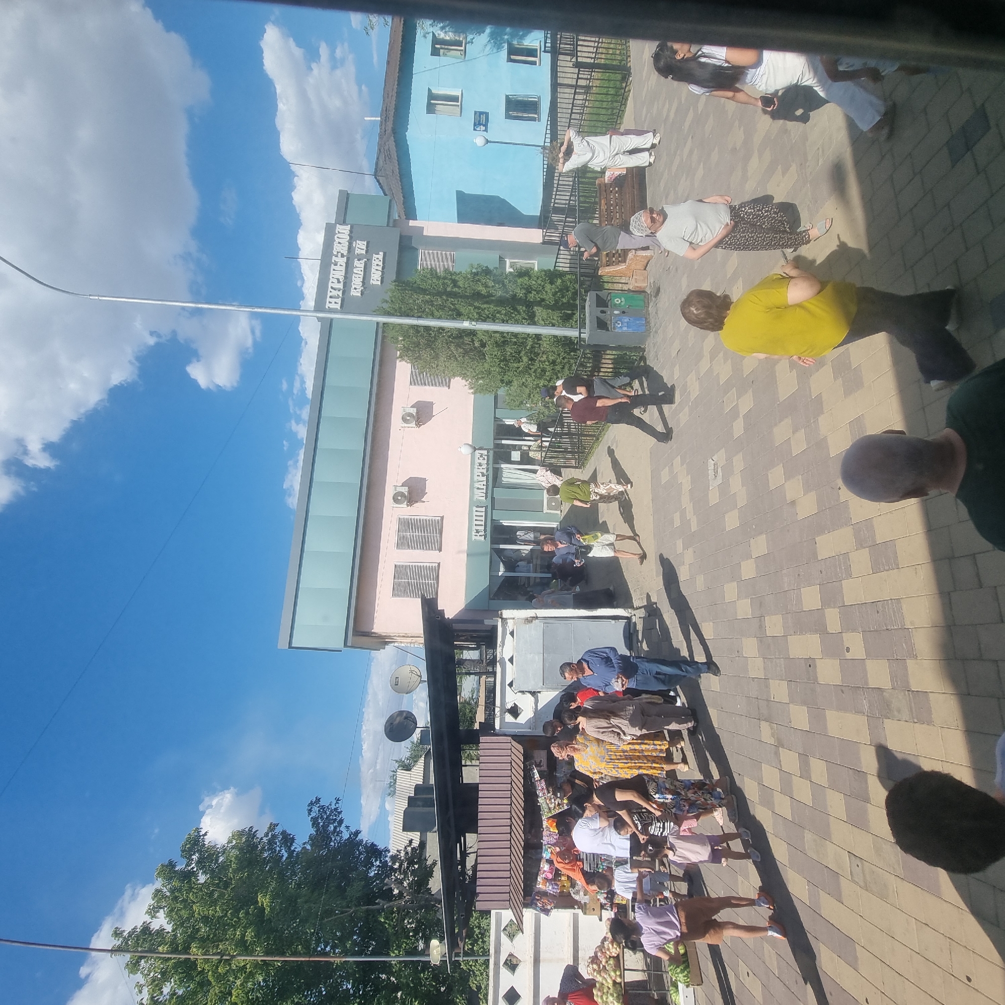 The image depicts a street scene in Turkistan, Kazakhstan, on a sunny day. A light blue building with white window frames is visible on the right, with a sign that appears to read "Nurly-Jol" and "Hotel". To its left is a pastel pink building with a green trim, labeled "KIPI MARKET". In front of these buildings, a paved area is populated with people. Several individuals are browsing goods displayed at a market stall on the left, where fruits and watermelons are visible. Other people are walking on the pavement or standing in groups. The sky is blue with scattered white clouds. The scene appears to be an outdoor marketplace and a commercial area in the city.
