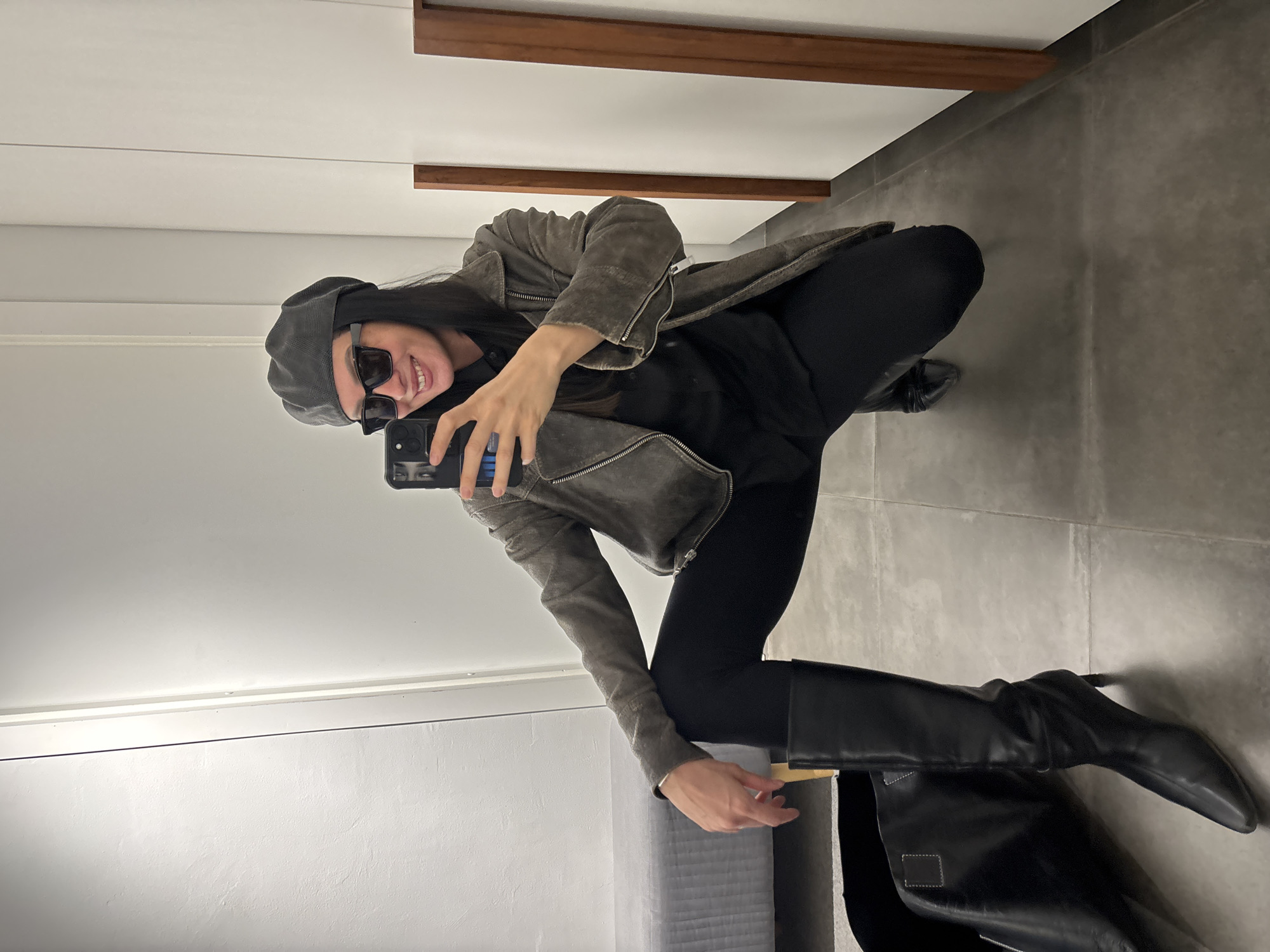 A woman is taking a selfie in what appears to be a fitting room. She is squatting and facing the mirror, holding her phone up. She is wearing sunglasses, a grey beret, a grey jacket, black leggings, and black knee-high heeled boots. A black bag rests by the boots. The fitting room has a grey floor, white walls, and wooden trim. There is no information available to confirm that the location is in the city of Chaikovsky, Russia.