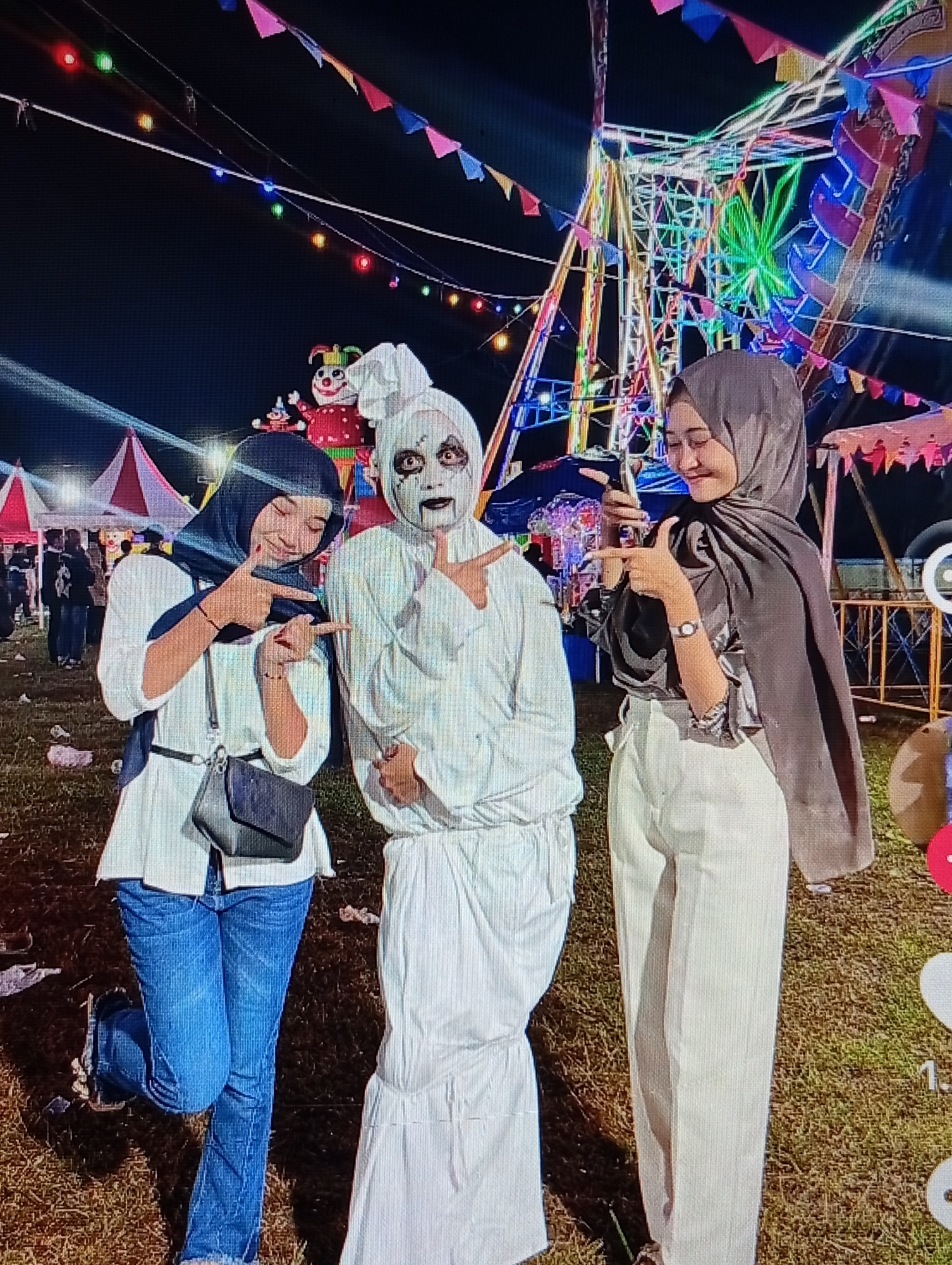 The image captures three individuals posing at a festive outdoor night event in Lhokseumawe, Indonesia. In the center stands a person in a white full-body costume, including a head covering, with white face paint and stark black eye and mouth makeup, making a forward-pointing gesture with both hands. To the left, a smiling woman wearing a dark hijab, a white long-sleeved top, blue jeans, and a dark cross-body bag makes a pointing gesture with both hands towards the central figure. To the right, another smiling woman in a grey hijab, a dark long-sleeved top, and light-colored trousers holds a smartphone in her right hand while giving a thumbs-up with her left.
The background features a large, brightly illuminated Ferris wheel with multi-colored lights. Above, strings of colorful pennant banners and individual decorative lights are strung across the dark sky. To the left and behind the subjects, several tents, some with red and white striped patterns, are visible, suggesting market stalls or booths. A large, inflatable red and white clown figure with a yellow collar is partially visible behind the central figure. Metal barriers are present on the right side of the frame. The ground is covered with grass and appears somewhat uneven, with minor debris. A partial user interface displaying social media-like heart icons and numerical text is visible in the bottom right corner of the image.