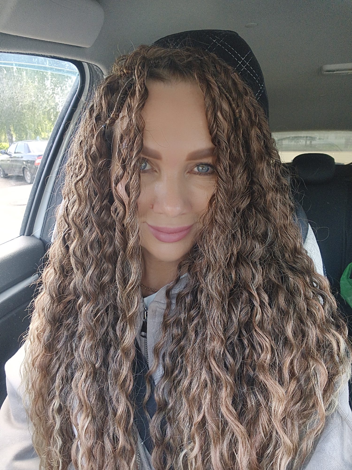 A close-up, eye-level portrait shows a woman seated in a car, likely in the driver's or front passenger seat, looking directly at the camera. She has long, voluminous, very curly and wavy brown hair with prominent lighter blonde or caramel highlights that frame her face and extend well past her shoulders. Her blue eyes gaze forward, and she has a subtle, pleasant smile on her lips, which are colored with light pink lipstick.
She is wearing a light-colored (possibly beige or grey) zipped-up top or jacket. Behind her head is a dark car headrest with a faint checkered pattern. Through the car window on the left, daylight illuminates the scene, and outside, green trees and a dark car are visible, suggesting it is daytime with clear weather. The interior of the car is generic. No visible text is present in the image.