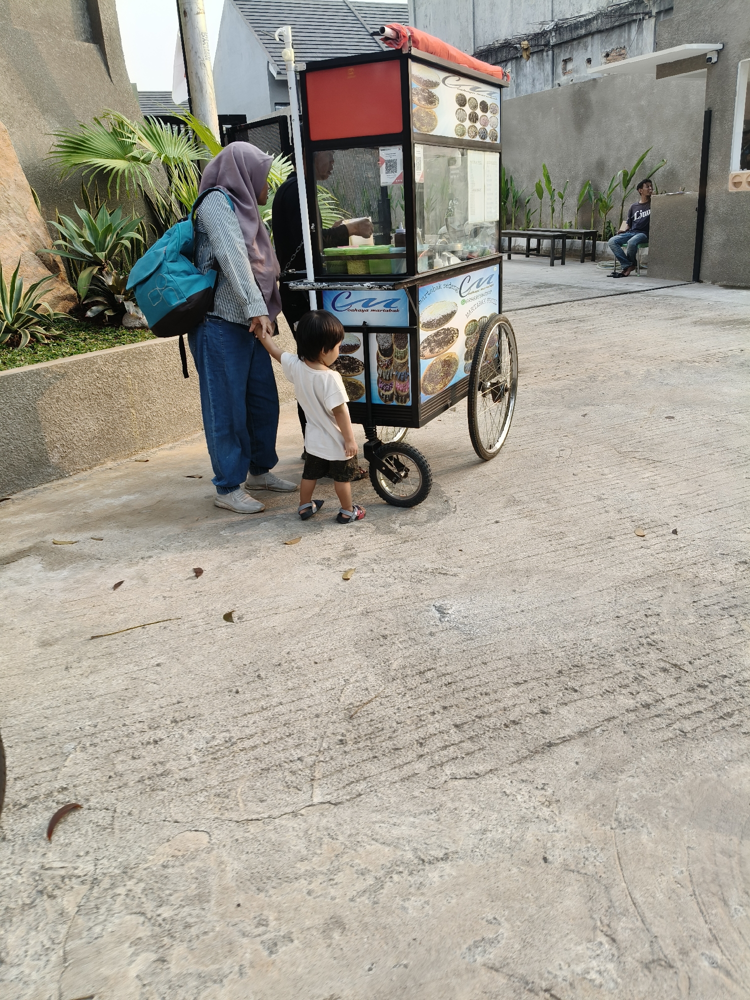 A woman in a grey hijab and a striped long-sleeved shirt, wearing blue jeans and white sneakers, stands holding hands with a young child in a white t-shirt and dark shorts. The child is wearing sandals. They are standing next to a food cart that displays pictures of food items and the text "Cahaya Martabak". The cart has a red top and a glass display case. Behind the woman and child, a man wearing a dark t-shirt and jeans is sitting on a chair, facing away from the camera. The scene appears to be outdoors, on a paved surface, with a concrete wall and some greenery in the background. This image was taken in South Tangerang, Indonesia, as indicated by the food cart's signage, which is a common type of street food vendor in the region.