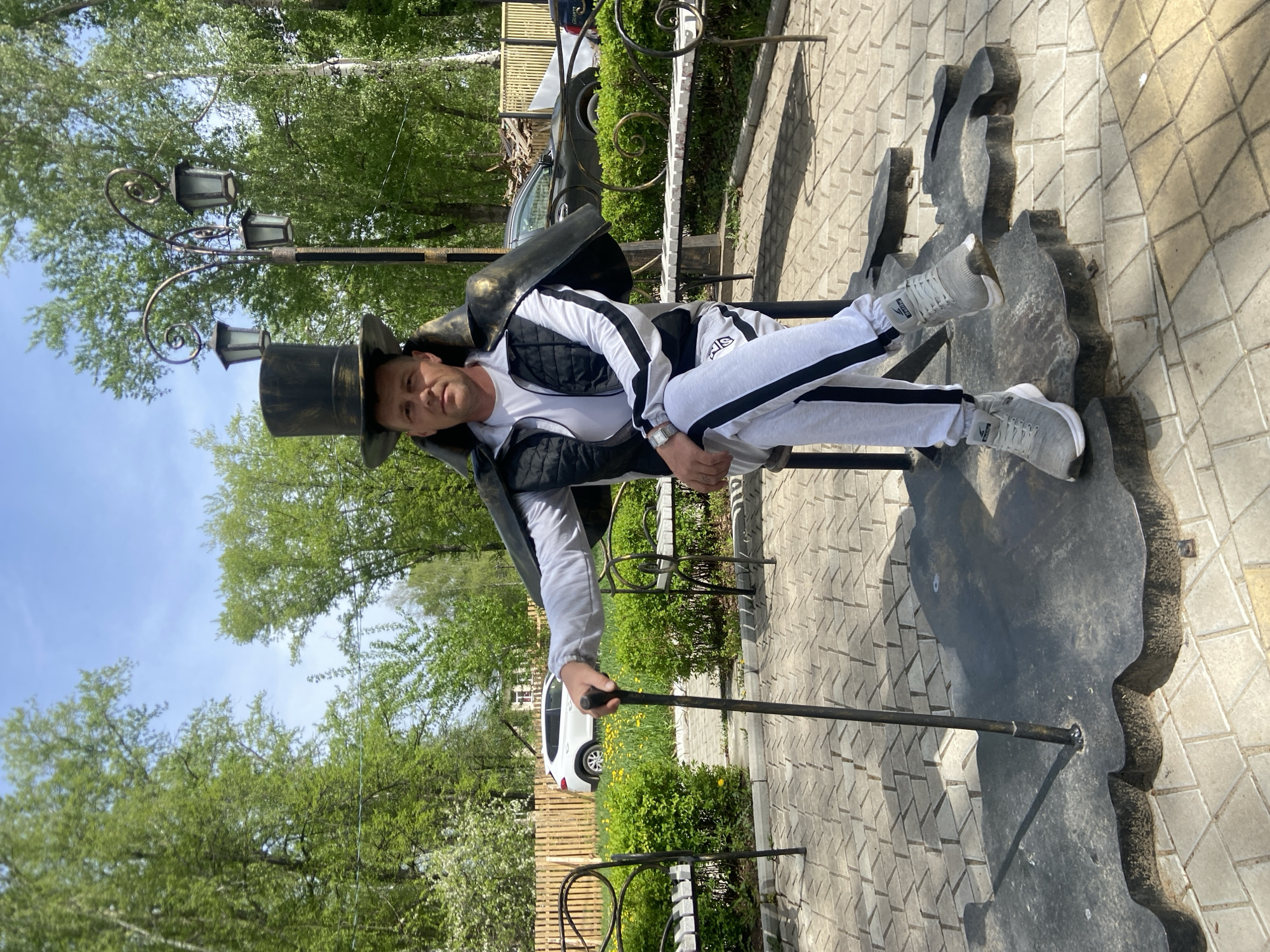 The image depicts a person in a white and black tracksuit, top hat, and holding a cane, leaning against a bronze sculpture on a paved area. The person wears light-colored sneakers. The sculpture is dark gray with rough edges. A lamppost with three lamps rises behind the sculpture. A fence, bushes, and trees are visible in the background, along with a white vehicle. The setting appears to be outdoors, possibly a park or public area in the city of Chaikovsky, Russia.