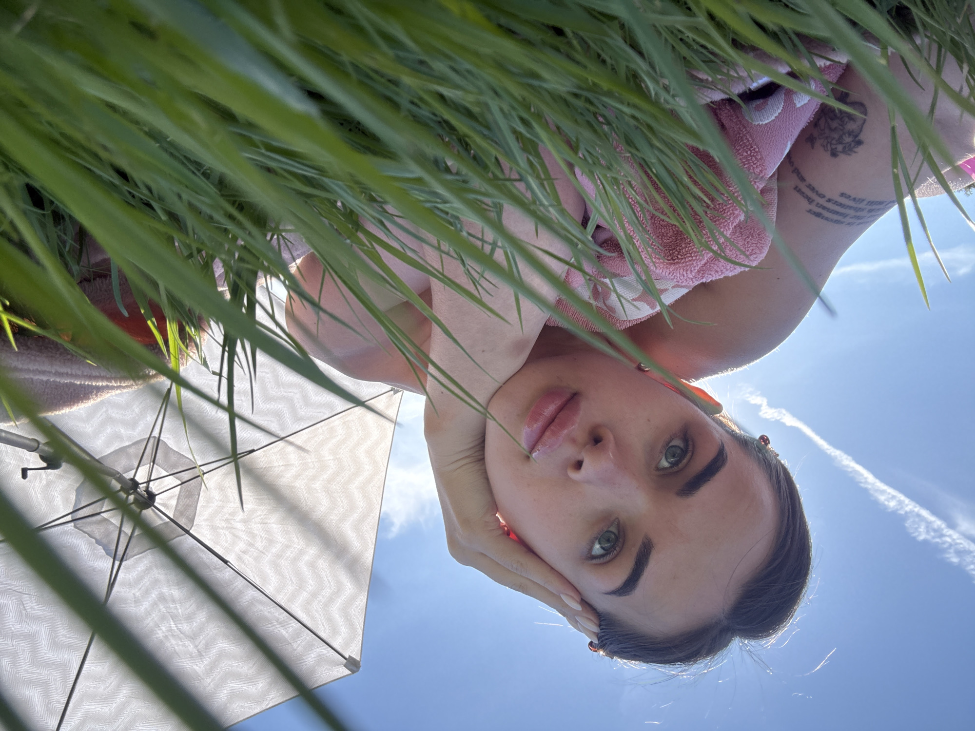 A low-angle shot of a woman lying down near grass and a white umbrella, looking up at the camera against a blue sky with a contrail. The location is unknown but potentially Лобаново, Russia. The woman rests her head on her hand. She is wearing a pink towel. Her arm shows tattoos.