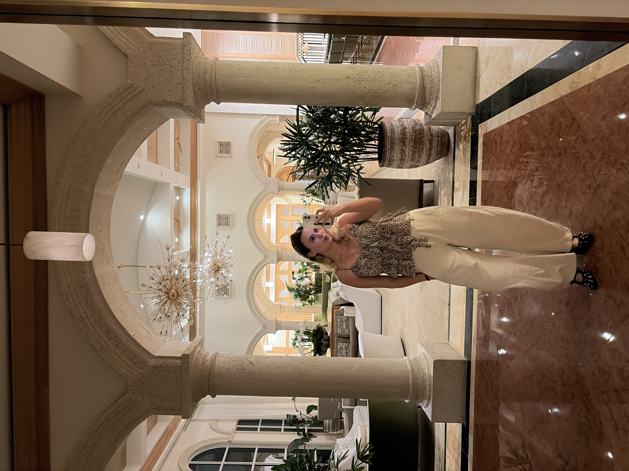 A woman is taking a selfie in an opulent lobby. She wears a patterned top and light-colored pants. The lobby features stone arches supported by pillars, ornate chandeliers, potted plants, and marble flooring. The location appears to be in Key Biscayne, United States of America.
