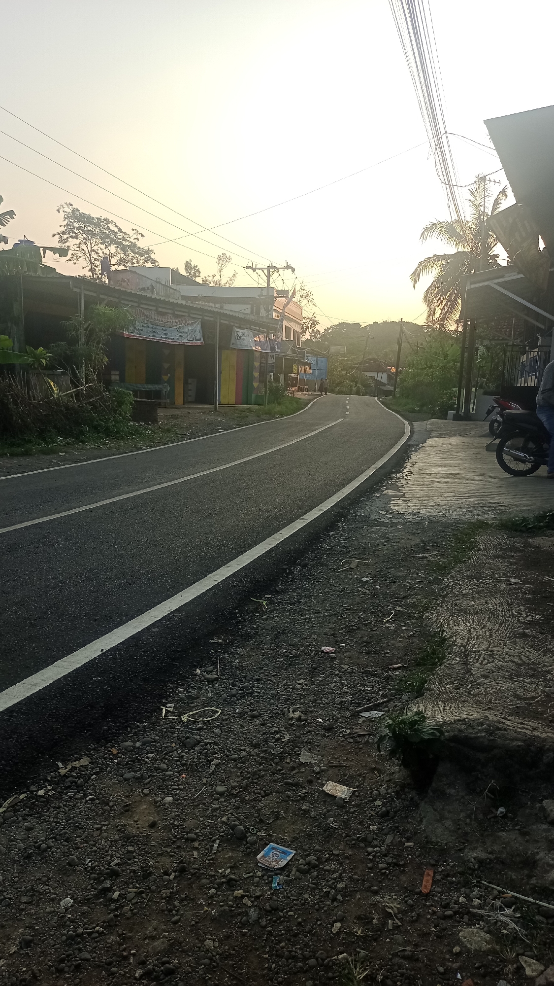 A paved road curves through a rural Indonesian setting near Cogreg. The sun is low in the sky, casting a warm glow and creating long shadows. On the left side of the road, several small buildings line the street, some with colorful facades. A few trees and dense vegetation are visible behind the buildings. Overhead, power lines stretch across the frame. On the right side of the road, a concrete sidewalk is wet, suggesting recent rain. A motorcycle is parked on the sidewalk. The road itself has clear white lane markings. The ground on either side of the road is covered in gravel and small stones, with some scattered litter visible, including a plastic wrapper and a piece of paper. In the distance, rolling hills can be seen under the hazy sky.