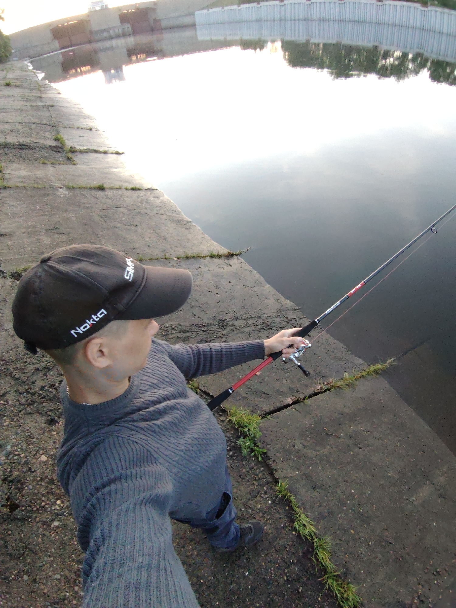 A person wearing a black baseball cap with the word "Nokta" on it, a gray ribbed sweater, and blue jeans stands on a concrete embankment holding a fishing rod. The rod is extended towards a body of water, likely the Kama River, in Chaikovsky, Russia. Cracks in the concrete are visible, with grass growing in them. In the background, there is a retaining wall, and a building can be seen in the distance, reflected in the water. The light suggests it is either early morning or late afternoon.