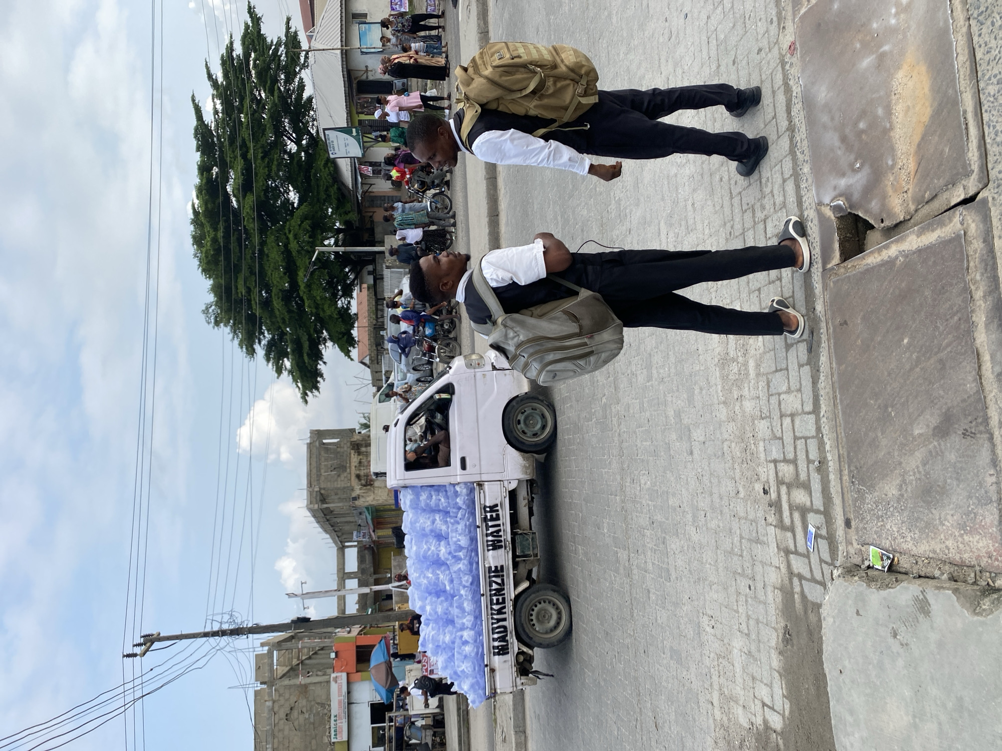 Two young men wearing white shirts, black vests, and black pants stand facing each other on a paved street, each with a backpack on. The man on the left has a grey backpack, and the man on the right has a tan backpack. Behind them, a light pink flatbed truck is loaded with numerous blue plastic water bottles. The truck has "MADYKENZIE WATER" written in black on its side. The background shows a street with other vehicles, buildings under construction, and trees. The sky is partly cloudy.