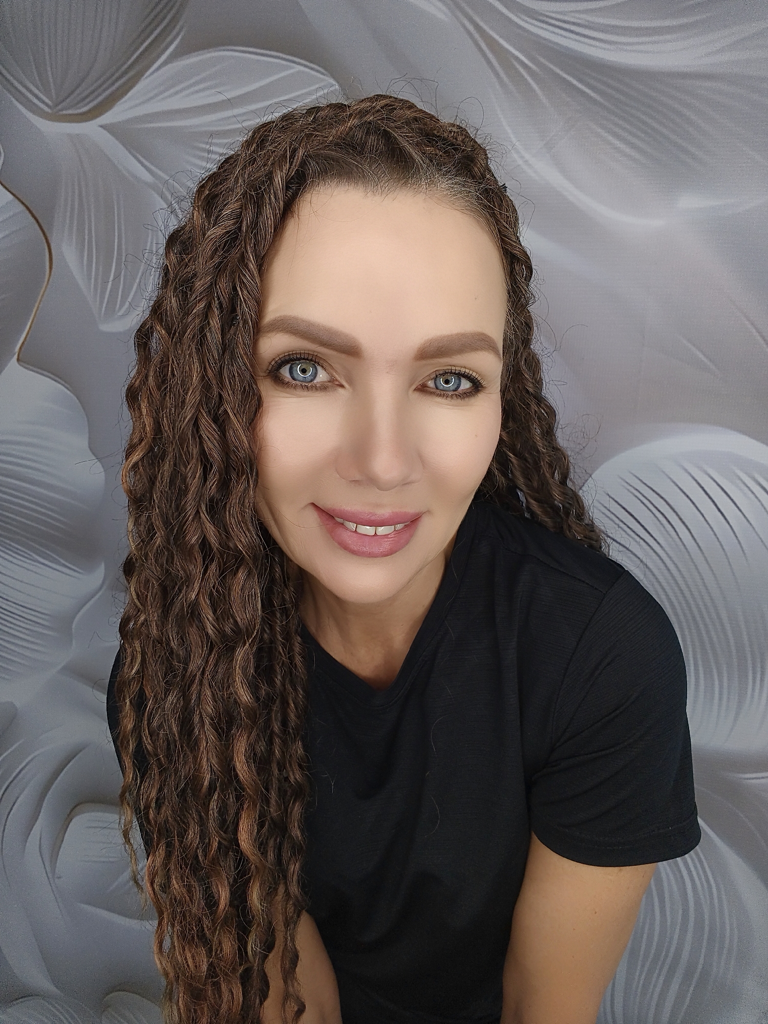 A medium close-up portrait shows a woman with long, curly brown hair, a black t-shirt, and bright blue eyes. She is smiling with her lips slightly parted, revealing her teeth. The background features a light gray backdrop with a white floral or feather-like pattern. The lighting is soft and even, highlighting the woman's features. The image appears to be taken indoors, possibly in a studio setting. The woman is positioned centrally in the frame, and her gaze is directed towards the camera.