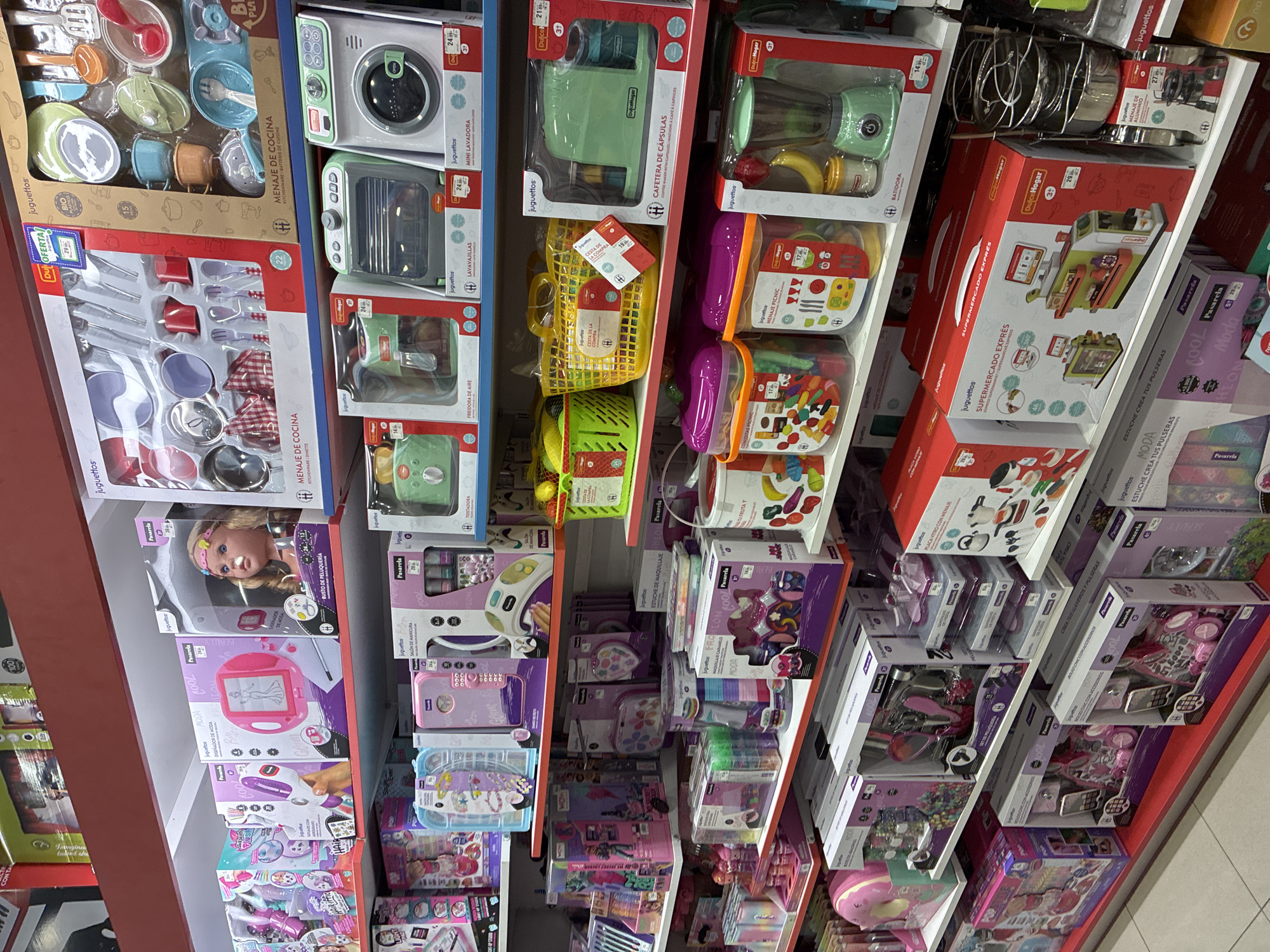 The image displays a well-lit retail store aisle, likely a toy store, in L'Hospitalet de Llobregat, Spain. The shelves are packed with colorful toy sets, predominantly for children. The toys appear to be categorized by play themes, with prominent displays of kitchenware sets, doll accessories, makeup kits, and kitchen appliances like blenders and washing machines. Many of the toy boxes feature Spanish text, indicating the target market. The overall environment is clean and organized, suggesting a typical retail display designed to attract customers. There are no people visible in the image.