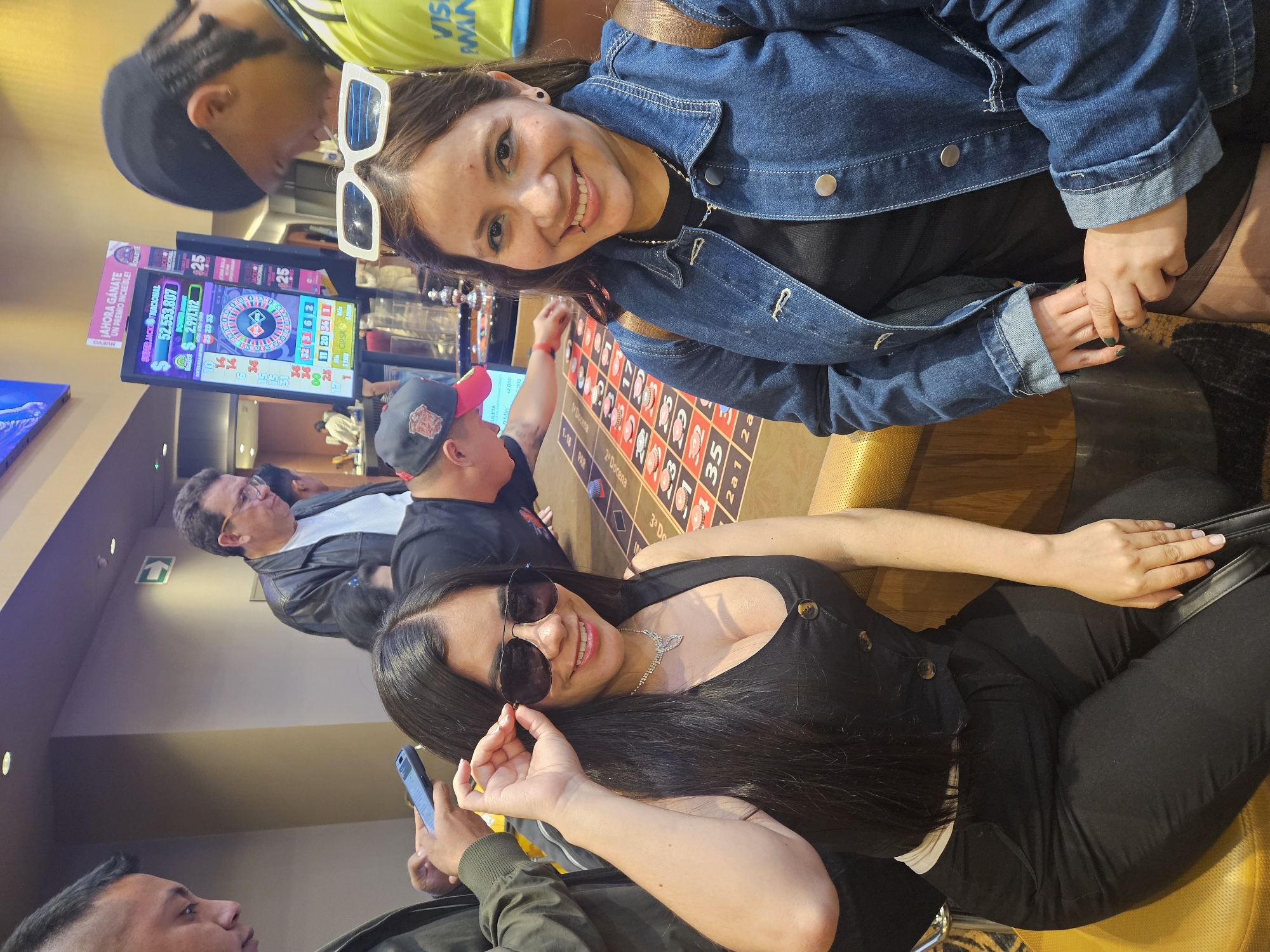 The image appears to have been taken inside a casino. Two women are posing for a photo at a roulette table. One woman is wearing a denim jacket and white sunglasses on her head. The other woman is wearing sunglasses and a black outfit with a silver necklace. Behind them, there are other people at the table and a large screen displaying gambling information. The OCR text indicates that it may be the Grand Casino in Bogota, Colombia.