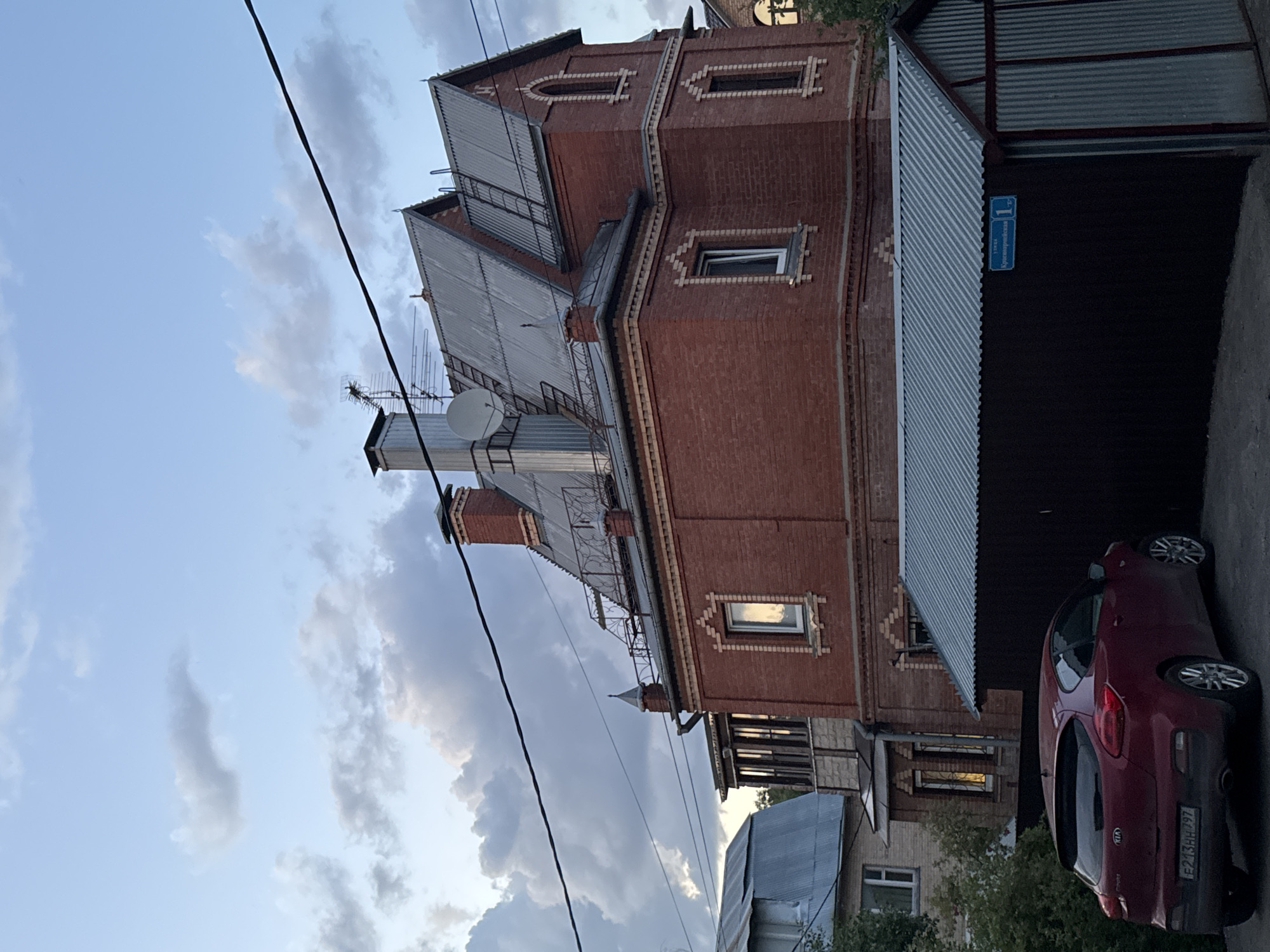 The image displays a street scene featuring a distinctive red brick building under a partly cloudy sky. The multi-story brick structure, positioned prominently, exhibits intricate architectural details, including decorative lighter brickwork around its rectangular and arched windows. Its complex roof is made of corrugated metal, supporting multiple chimneys, a small balcony with a metal railing, a satellite dish, and a television antenna.
Adjacent to the brick building, to the right, is a garage or shed with a corrugated metal roof. A blue street sign is visible on its wall, bearing the Cyrillic inscription "ул. Сумская" (ul. Sumskaya), indicating "Sumskaya Street," followed by the building number "11." A red Kia car is parked in front of this structure, with its rear facing the viewer. The license plate "Е213НН 797" is clearly legible.
Overhead, dark power lines traverse the sky, which features scattered clouds and hints of a brighter, lighter blue beyond. The lighting suggests it is either late afternoon or early evening. Patches of green foliage are visible towards the bottom right, indicating the presence of trees or shrubs in the vicinity. The scene depicts a quiet residential area.