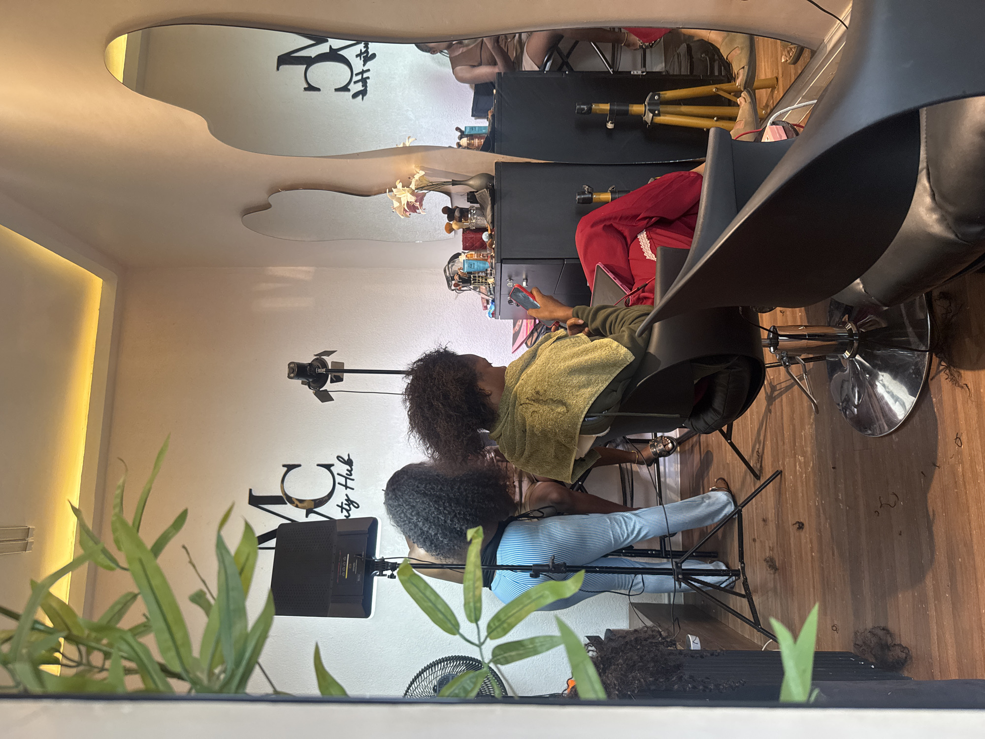 The image depicts the interior of a beauty salon in Abuja, Nigeria. The scene is viewed through a reflective surface, possibly a window or a mirror. In the foreground, a person with dark, curly hair is seated in a black salon chair, wearing a green towel around their shoulders and red pants. They are looking down at a smartphone in their hand. To the left, another person with long, dark, curly hair, wearing blue and white striped pants, is standing and appears to be working on the hair of the seated individual or another unseen person. A professional ring light is positioned behind the standing individual, illuminating the workspace.
The salon features light-colored walls with a large, stylized "MC Beauty Hub" logo prominently displayed. Two large, irregularly shaped mirrors hang on the wall, reflecting parts of the salon. A black cabinet with various beauty products and brushes is visible against the wall. The floor is made of wood and shows scattered strands of hair and small clippings, indicating recent hair services. Other equipment, including a tripod and a fan, are also present. The lighting appears to be a combination of overhead illumination and professional salon lighting. The overall impression is a functional and stylish beauty salon environment.