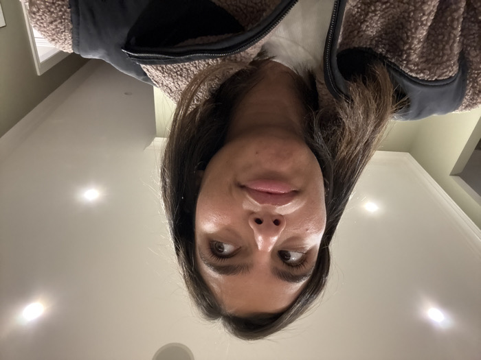 The image depicts an indoor scene focused on a young adult female. She has dark brown hair, brown eyes, and is looking upwards and to the right with a slightly pursed mouth, conveying a contemplative or pensive expression. She is wearing a light-colored (likely white) long-sleeved top underneath a brown, fuzzy sherpa-style vest or jacket with black fabric panels on the shoulders and collar. The background features a light-colored ceiling with multiple illuminated circular recessed lights. White crown molding runs along the top of a muted green or olive-colored wall. A partially visible doorway or opening to another room is present in the background to the right. The environment suggests a residential or office interior. No specific identifying markers for the city of Princeton, United States are visible in the image.