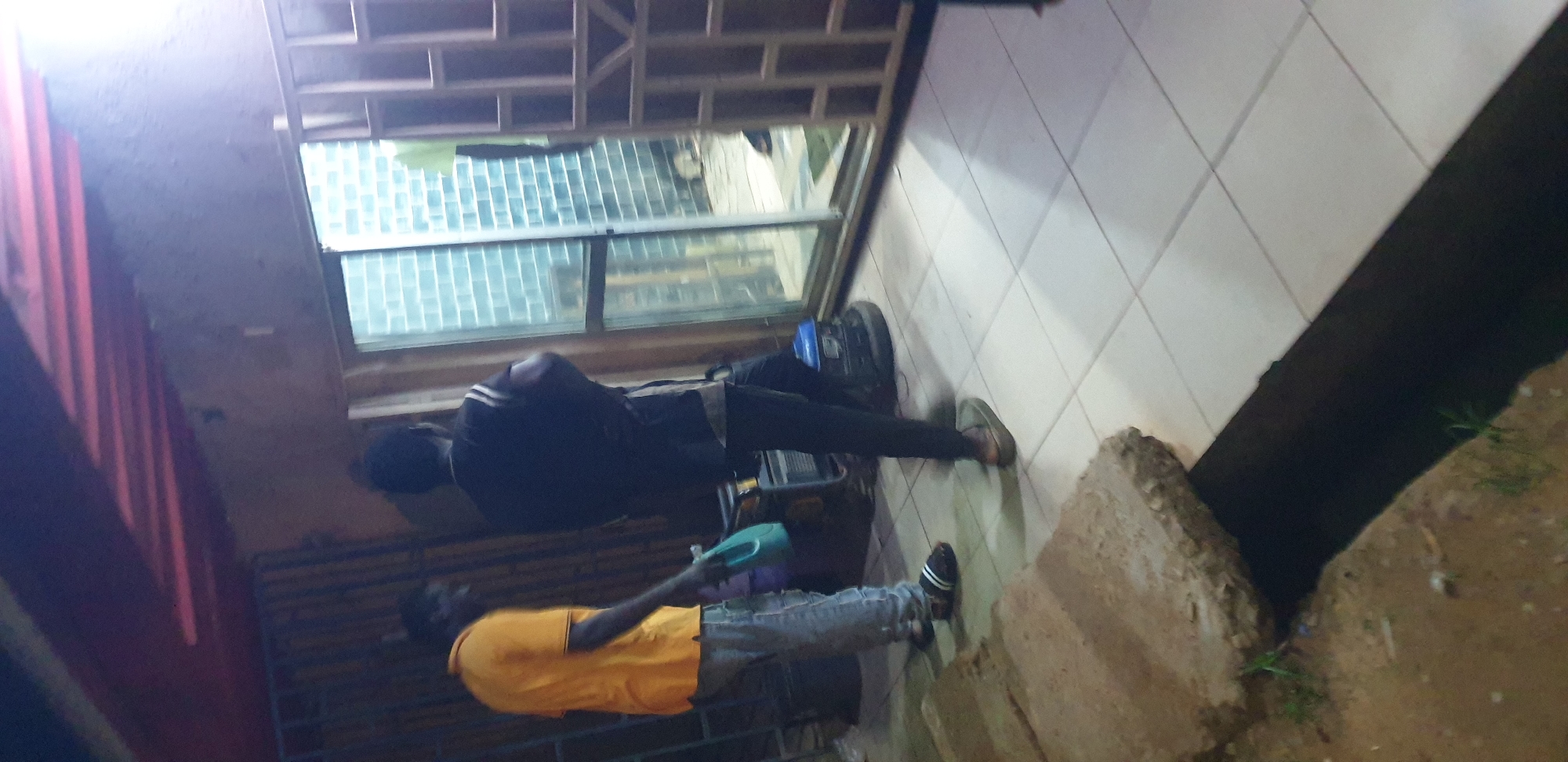 The image displays an outdoor or semi-enclosed area adjacent to a building in Narayi, Nigeria, under artificial illumination, indicating evening or nighttime.
Three individuals are present. In the left-midground, a male wears a yellow short-sleeved polo shirt, light-colored shorts, and dark slide sandals with white stripes. He holds a light blue plastic container with a handle in his left hand. To his right and slightly behind him, a second male is observed from the rear, attired in a dark sleeveless top with white shoulder stripes and dark pants. He is positioned facing a large glass sliding door or window. A third individual, partially visible through the glass to the far right, is wearing a light-colored (possibly green or yellow) short-sleeved shirt.
The ground in the immediate foreground consists of a dark, open drainage ditch running horizontally across the lower portion of the frame. To the left of the ditch, two large, irregularly shaped concrete slabs rest on dark brown earth sparsely covered with green vegetation. To the right of the ditch, the ground transitions to a surface tiled with light-colored (likely white or off-white) square ceramic tiles, extending towards the building.
Between the first two individuals, two pieces of machinery are visible. A larger, grey-colored unit, possibly a generator or pump, is situated behind the male on the left. To its right, a smaller, blue-colored machine, potentially another generator or a floor polisher, rests on a circular base.
The adjacent building features a light-colored wall. A prominent glass sliding door or window, divided into multiple panels, reveals an interior with a light blue brick wall and the partially visible third individual. To the far right, a light-colored security gate or grille with a geometric grid pattern is partially open. Overhead, a reddish-brown structure, consistent with exposed roof beams or rafters, suggests a covered overhang or porch area. The scene is characterized by functional elements and artificial lighting.