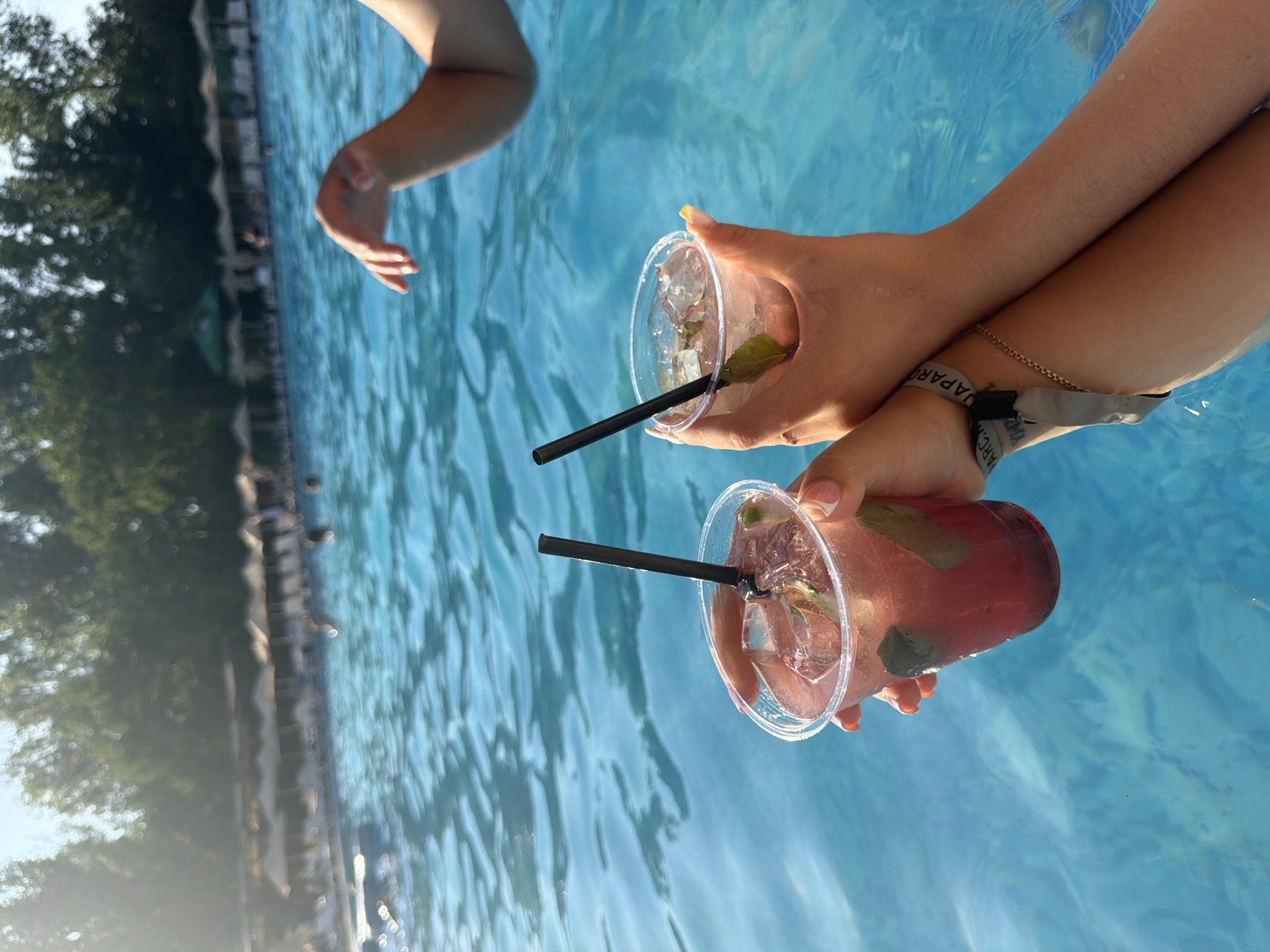 Two people are in a swimming pool, holding two red cocktails with ice and mint. The pool water is blue and appears to be outdoors, with a line of lounge chairs and umbrellas visible in the background. The sun is shining, casting a warm light on the scene. The individuals are wearing wristbands, suggesting they are at a resort or event. The city of Sociteni, Moldova, is noted as the location.