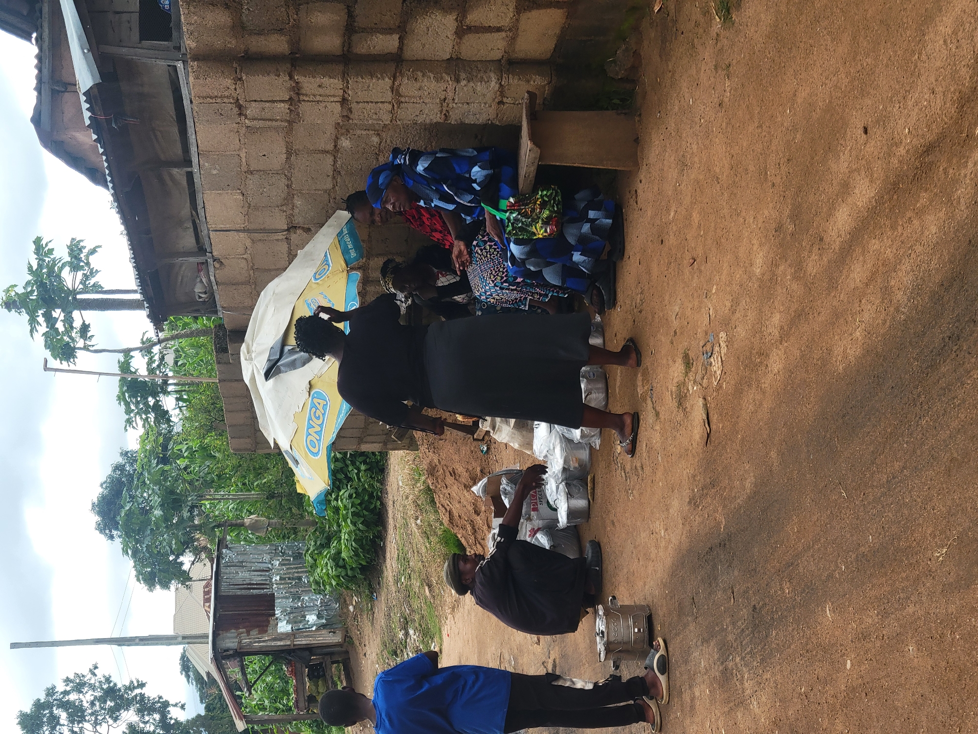 The image depicts an outdoor scene in what appears to be a market or roadside stall area in Jos, Nigeria. The ground is primarily composed of reddish-brown dirt with some scattered debris. On the left, a young person in a blue t-shirt and black pants stands facing away from the camera, near a silver portable stove. In the center, a man in a dark outfit and a green cap is crouched down, surrounded by several silver pots and packaged items. He is looking towards the right. To the right, a group of women are seated on a wooden bench against a rough brick wall. One woman, wearing a blue and black patterned outfit and headscarf, is looking to the left. Next to her, another woman in a colorful patterned dress is seated. A third woman, partially obscured, is also seated with them. Above the group, a yellow and white umbrella with the "ONGA" logo is providing shade. Behind the women, the brick wall extends upwards and is topped with a corrugated metal roof structure. Vegetation, including trees and corn stalks, is visible in the background, along with some rudimentary structures that suggest a built environment. The overall impression is of a casual, everyday commercial activity taking place in a somewhat informal setting.