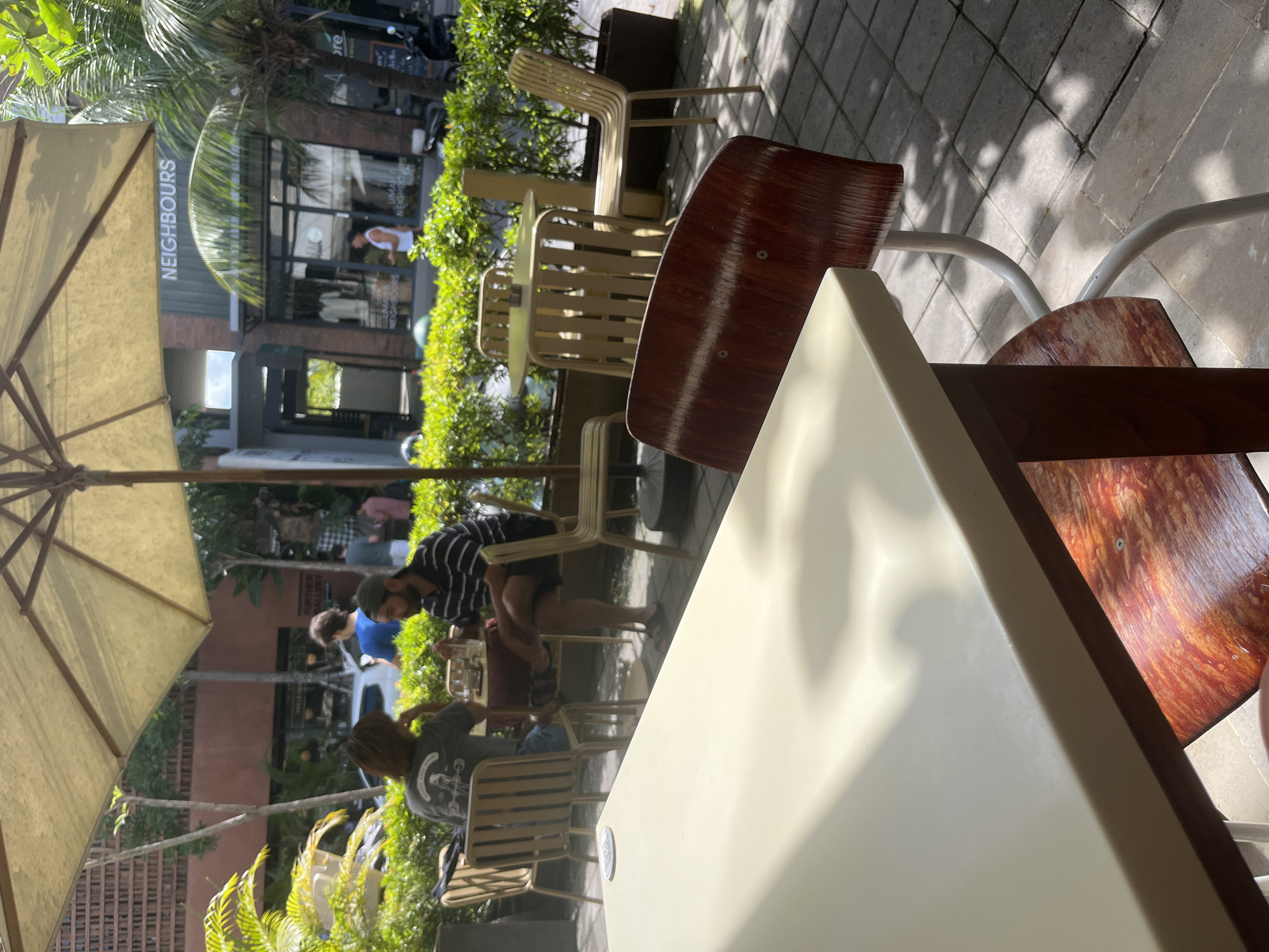 The image depicts an outdoor cafe scene in Mengwi, Indonesia. The foreground consists of cafe furniture, including tables and chairs. A partially visible umbrella shades the area. In the background, there's a building with a storefront labeled "NEIGHBOURS." People are seated at the cafe tables, engaged in activities such as eating and drinking. Lush greenery surrounds the seating area. The cafe appears to be located on a paved street or sidewalk.