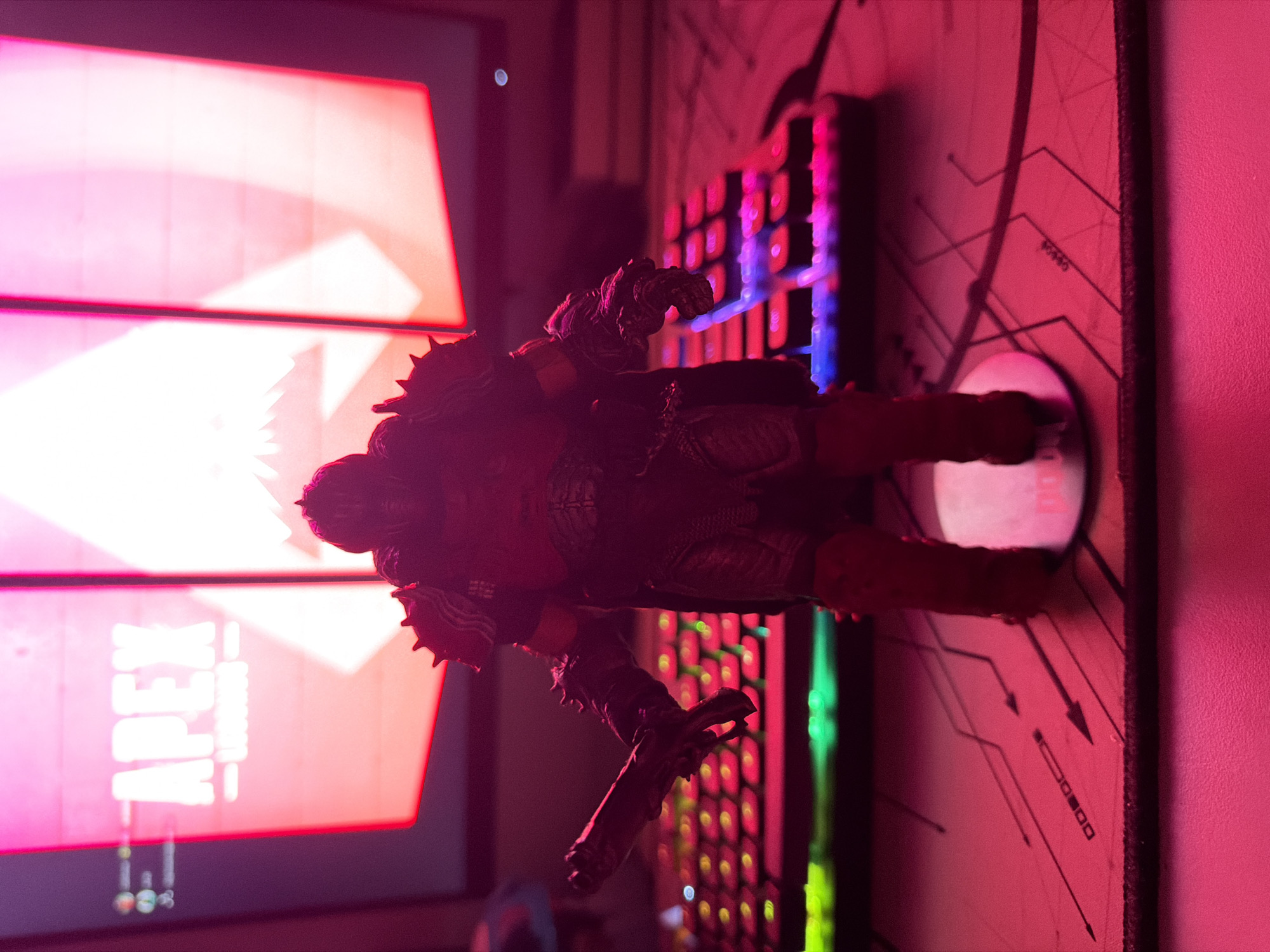 A red-lit gaming setup featuring a computer monitor displaying the game "Apex Legends", a figurine of a character with spiky armor on top of a colorful backlit keyboard, and a mousepad with circuit-like designs. The scene is indoors, under artificial lighting. There's no indication of the location being Belgrade, Serbia.
