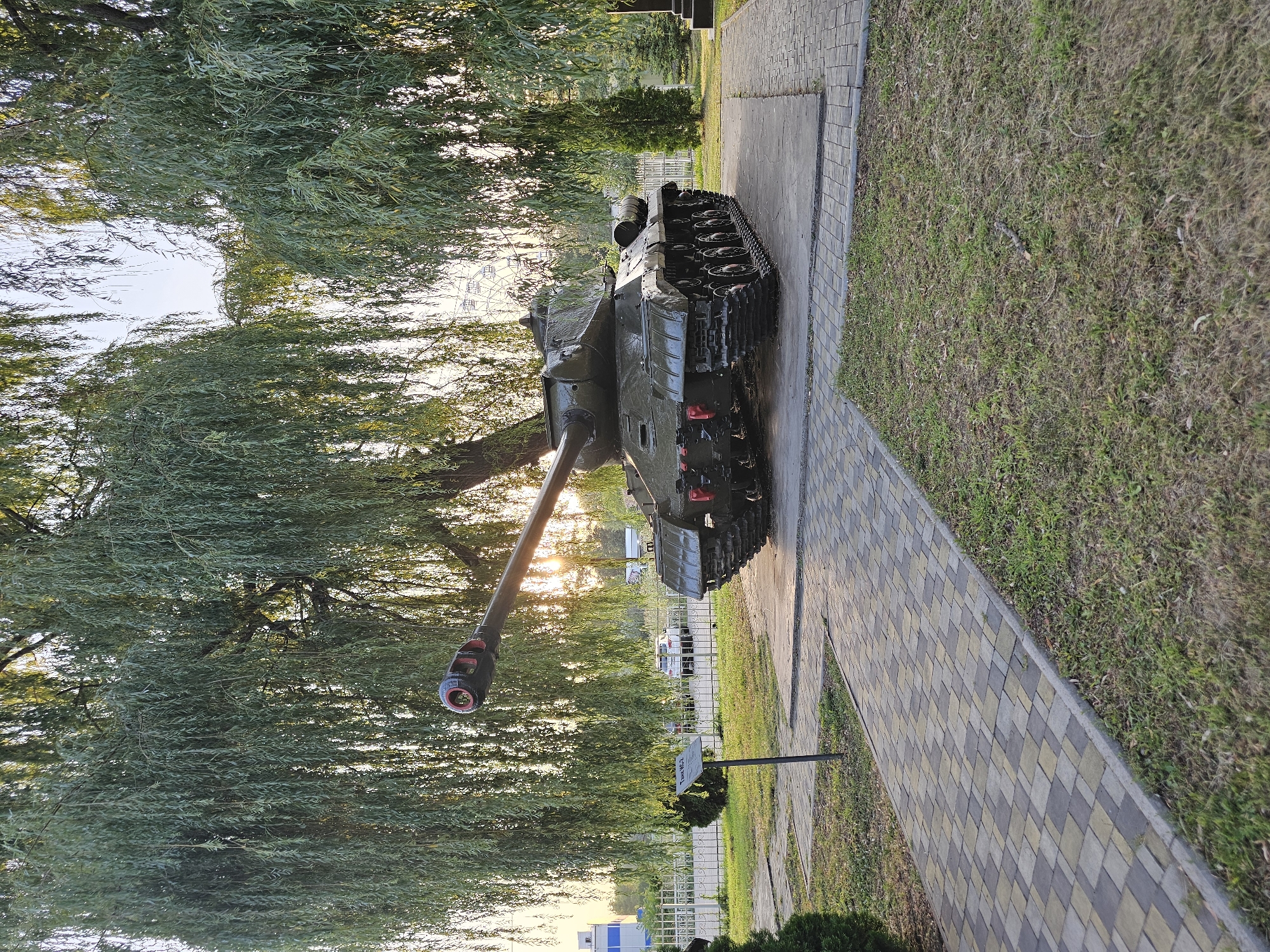 A military tank, identified as a IS-3 heavy tank, is displayed outdoors on a paved platform at the Alley of Heroes in Cherkessk, Russia. The tank is painted dark green and is positioned on a concrete slab surrounded by a paver walkway. Lush green trees with drooping branches, likely willow trees, form a canopy over the scene, with sunlight filtering through them. In the background, a fence, a white car, a building, and a Ferris wheel are visible. A signpost with text is placed near the tank. The overall environment suggests a public memorial or park setting.