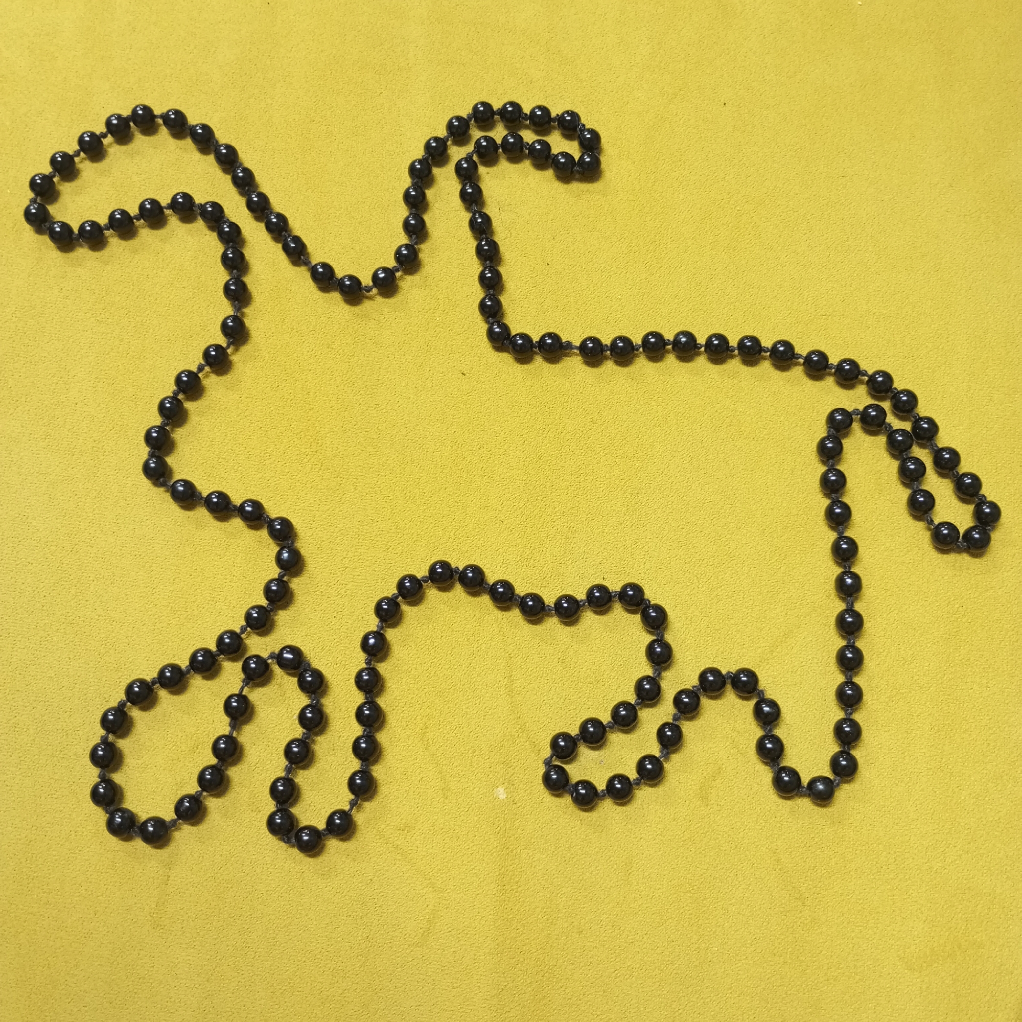 A black beaded necklace is arranged in the shape of a stylized, somewhat abstract horse silhouette against a mustard yellow background. The necklace outlines the head, neck, body, and legs of the horse. The background appears to be a solid, textured surface. The image is composed in a simple, straightforward manner. There are no people in the image. The city of Pervomaysky, Russia, is not relevant to the described scene.