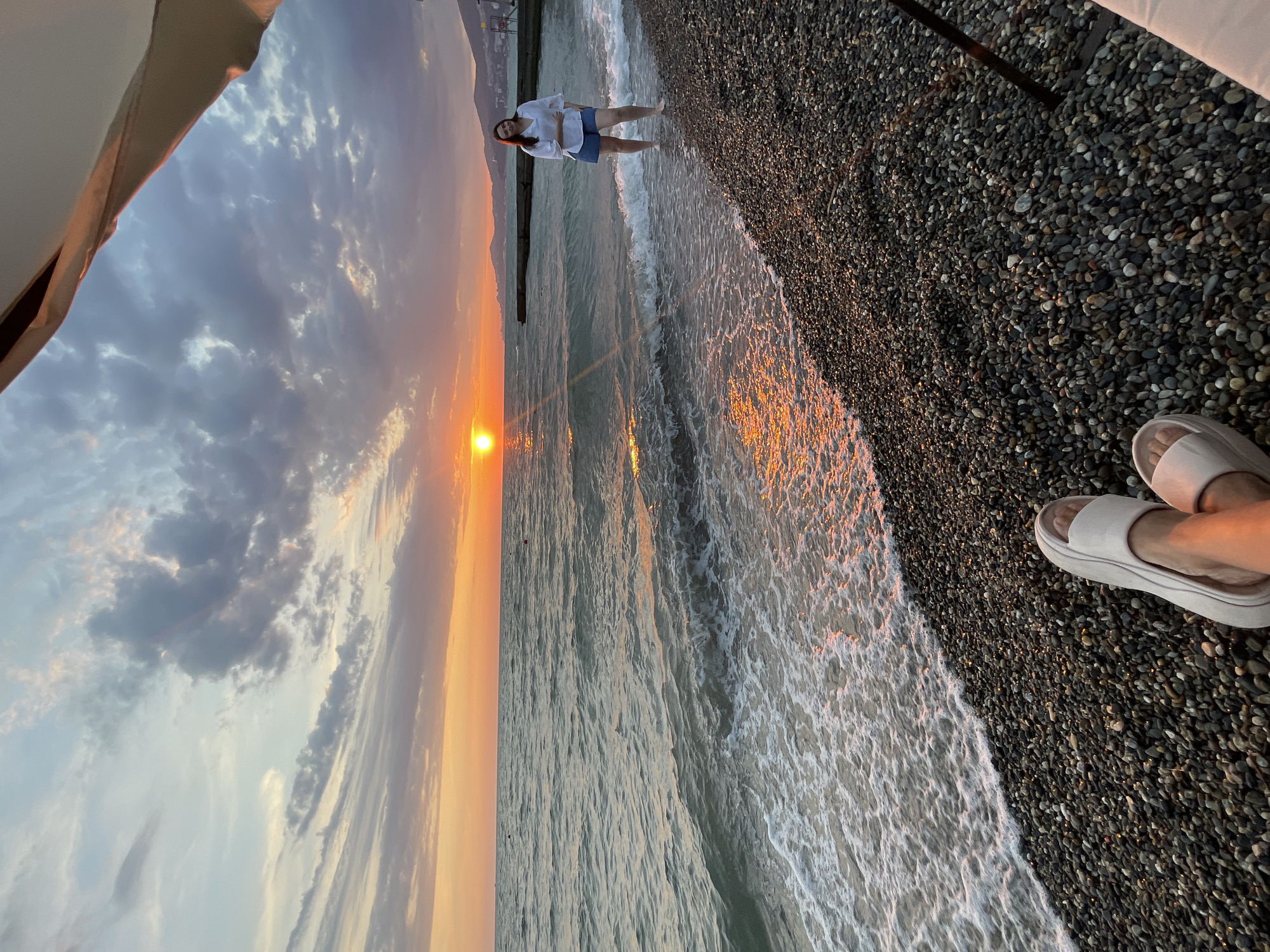 A woman stands on a pebble beach in Khostinskiy, Russia, near the Black Sea. The sun is setting, casting a warm orange glow over the water and sky. The waves are gently lapping at the shore, and the woman is looking towards the sunset. In the foreground, a pair of white sandals are visible, suggesting someone is sitting on the beach. The overall scene is peaceful and serene, capturing the beauty of a sunset over the sea.