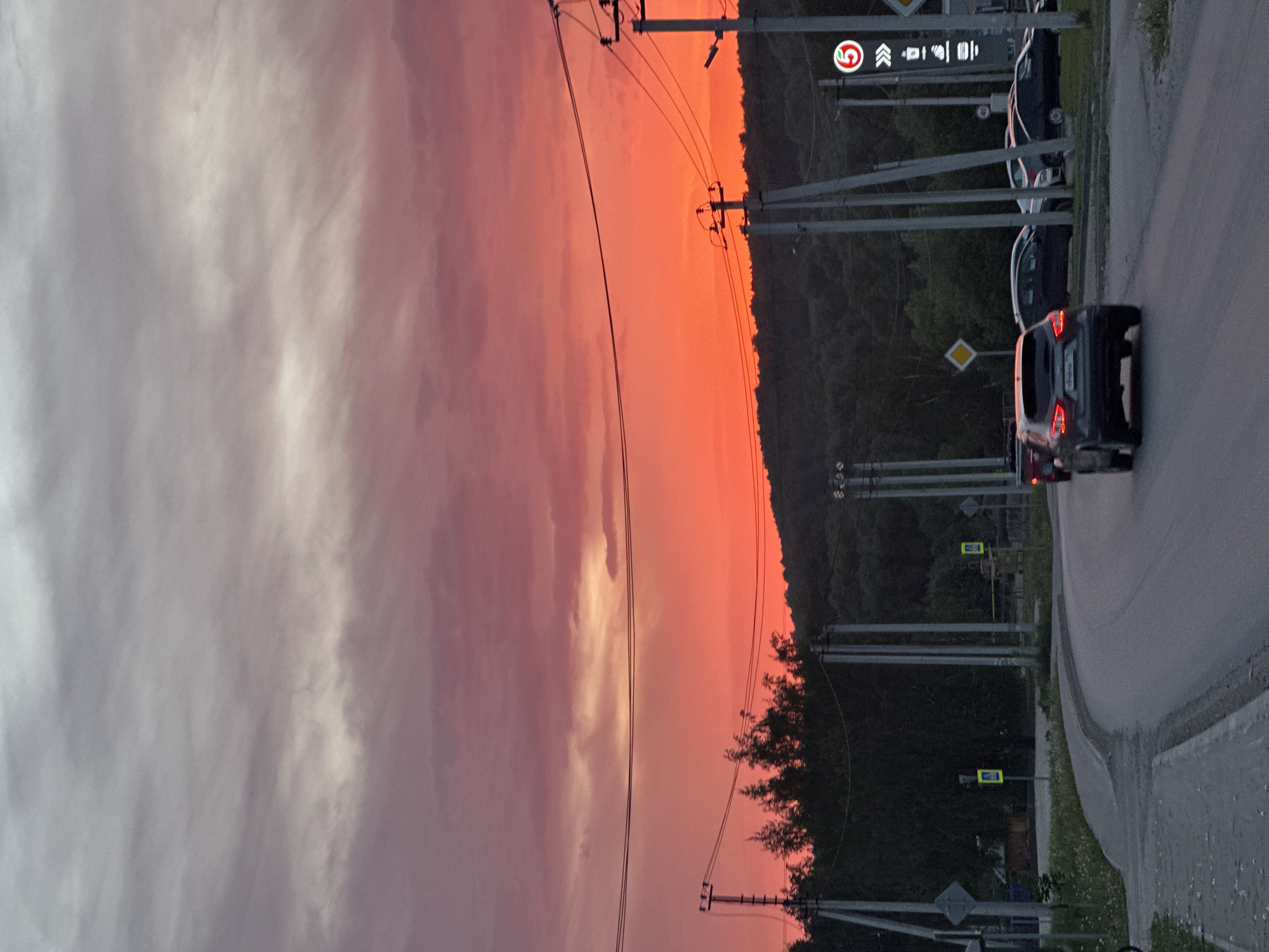 The image shows a street scene in Эстачи, Russia, at dusk. The sky is a mix of orange and pink, indicating a sunset. Power lines run across the upper portion of the image. A road runs along the right side with several parked or moving vehicles, including a dark-colored SUV with illuminated taillights. Road signs and pedestrian crossing markers are visible along the side of the road. Trees and vegetation line the backdrop of the street. A building marked with a "5" logo and other symbols is noticeable in the background.