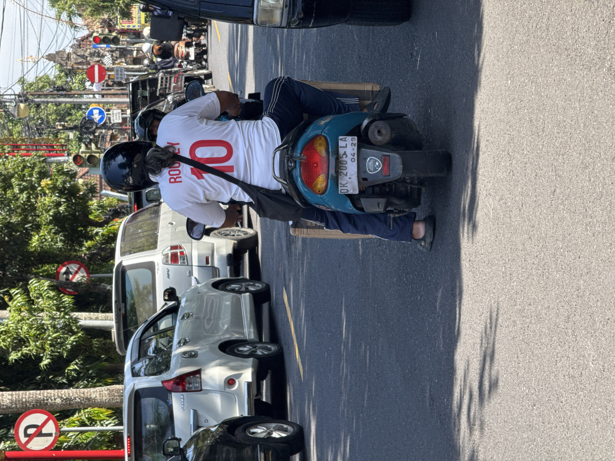 The image captures a street scene in Gianyar, Indonesia, during daylight hours, likely mid-morning or afternoon given the harsh shadows. The foreground is dominated by a person riding a teal-colored scooter. The rider, wearing a black helmet and a white t-shirt with red lettering and the number "10" on the back, is seen from behind. They are also wearing dark blue pants with white stripes. A black shoulder bag is slung across their chest. The scooter's license plate is visible and reads "DK 2005 LA" with a date "04-29" below it. Cardboard boxes are strapped to the rear of the scooter.
The scooter is in traffic, surrounded by other vehicles. To the left, a white car is partially visible. Behind the scooter, other motorcycles and cars are present, suggesting a busy urban environment. Traffic lights and street signs are visible in the background, indicating an intersection or main road. Trees line the street, their foliage creating dappled shadows on the asphalt. The overall impression is of a typical traffic scene in a bustling Indonesian city.