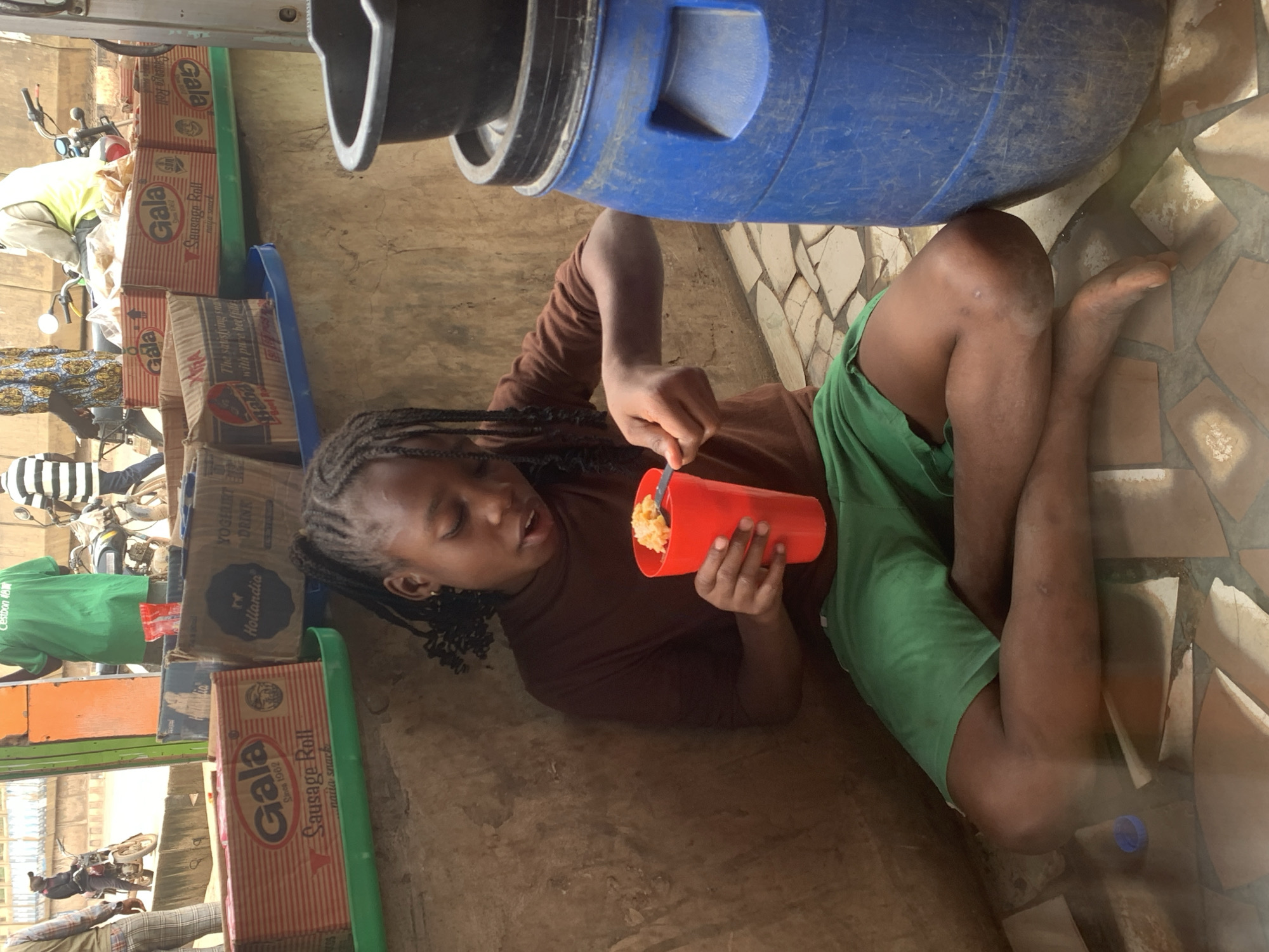 A young Black girl with braided hair sits on the ground in Shagamu, Nigeria, eating from a red cup with a fork. She wears a brown long-sleeved shirt and green shorts. In front of her are several cardboard boxes of Gala sausage rolls and Hollandia yoghurt drinks, some stacked on a green tray. To her right is a large blue plastic barrel, and a black container rests on top of it. In the background, people are visible on motorcycles, and one person wears a green shirt with "Cestbon RGM" printed on it. The ground is paved with irregular, broken tiles. The scene suggests a bustling market or roadside stall environment.