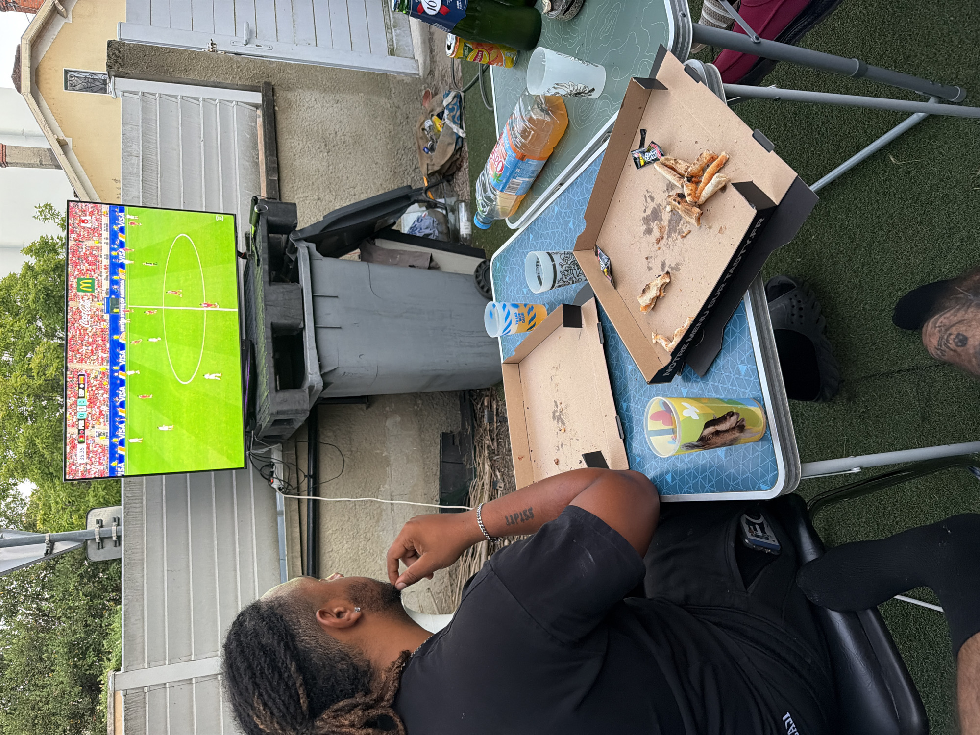 A medium shot captures an outdoor scene in Villeurbanne, France, where two individuals appear to be watching a soccer match on a large television screen. The television is positioned on a plastic stand in front of a grey garbage bin. In the foreground, a person with dark dreadlocks is seated, facing away from the camera, wearing a black t-shirt and black pants. Their left arm, tattooed with the word "LUPISS," rests on their chin, and they are wearing a silver bracelet. A second person's legs, clad in black socks and patterned pants, are visible in the lower right portion of the frame.
Two folding tables are set up. The closer table is blue and features two empty pizza boxes, one with a few remaining crusts and packets of sauce. Several disposable cups and a can of soda are also present on this table. The further table, green, holds a large bottle of orange soda, a green bottle with a red label, and additional disposable cups. The background reveals a residential building with a yellow facade and a white fence. The setting appears to be a backyard or patio with artificial turf flooring.