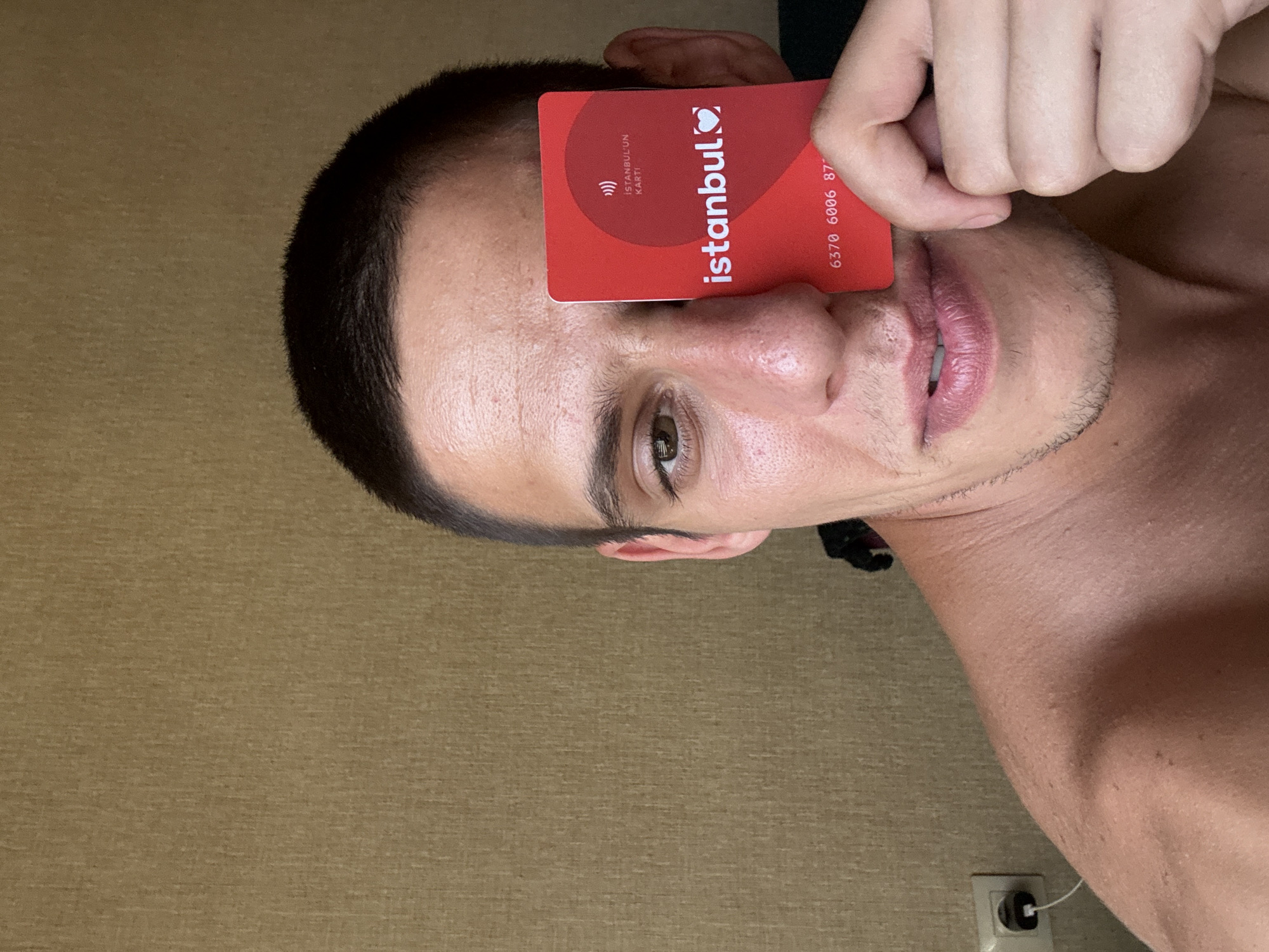 A man is holding a red Istanbulkart in front of his right eye, obscuring it. The card has white text and logos, including "ISTANBUL'UN KARTI" at the top, a heart logo with "istanbul" inside, and a series of numbers "6370 6006 87" at the bottom. The man has short dark hair and is looking directly at the camera. He is shirtless and appears to be indoors, with a textured brown wall as the background. A white electrical outlet with a plug is visible on the left side of the image. The scene does not contain any elements indicating it is in Sofia, Bulgaria.