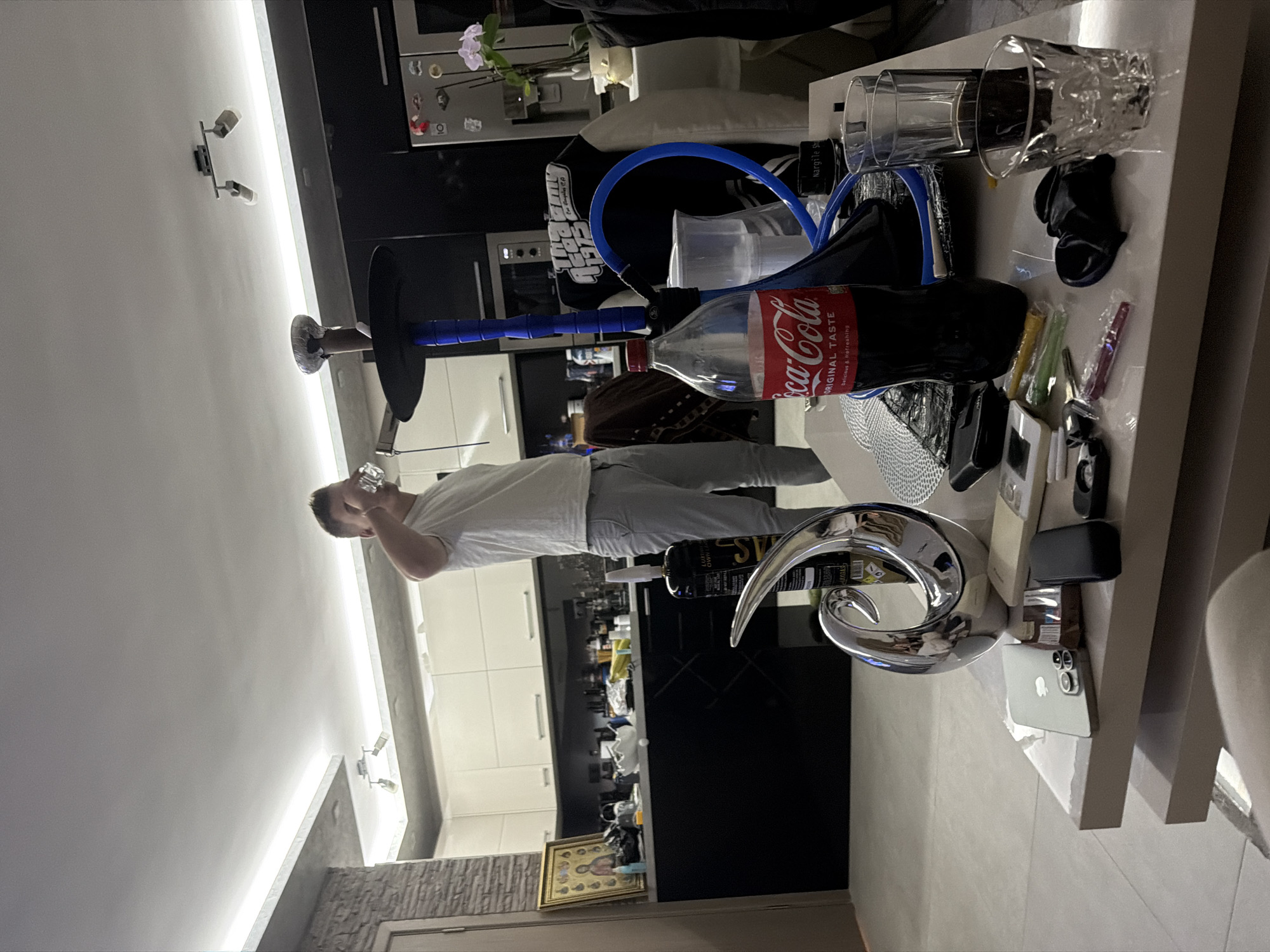 An indoor scene depicts a modern living space with an open-plan kitchen, viewed from a slightly elevated perspective. A man, dressed in a white t-shirt and grey pants, stands on the left side of the frame, drinking from a clear glass. His face is partially obscured by the glass.
In the foreground, a white coffee table is cluttered with various items. Prominently featured is a black Coca-Cola bottle, which appears to be mostly empty, and part of a blue hookah pipe with a black base. Several clear drinking glasses, including one with a textured pattern, are also on the table. Other items include a black, abstract decorative object, an iPhone (likely a Pro model) lying face-down, a chrome-colored abstract sculpture, small wrapped plastic tips (yellow, green, pink), and a small black case or device. A black fabric item, possibly a t-shirt, bearing a "Grand Theft Auto" logo and the numbers "1975" is visible near the hookah base. Text labels such as "Nargile S" and "LUXURY OWN L" are also discernible on various objects.
The background reveals a contemporary kitchen area with sleek black base cabinets and white upper cabinets, white countertops, and a built-in oven or microwave. An orchid plant in a pot sits on a counter, and a refrigerator with magnets is partially visible. The ceiling features modern strip lighting and two small spotlight fixtures. A framed religious icon or painting leans against a lower shelf in the kitchen area. The walls are a mix of light-colored plaster and a textured stone-like accent. The overall impression is a casual, relaxed setting, likely an apartment or house, possibly in the evening given the reliance on artificial lighting.