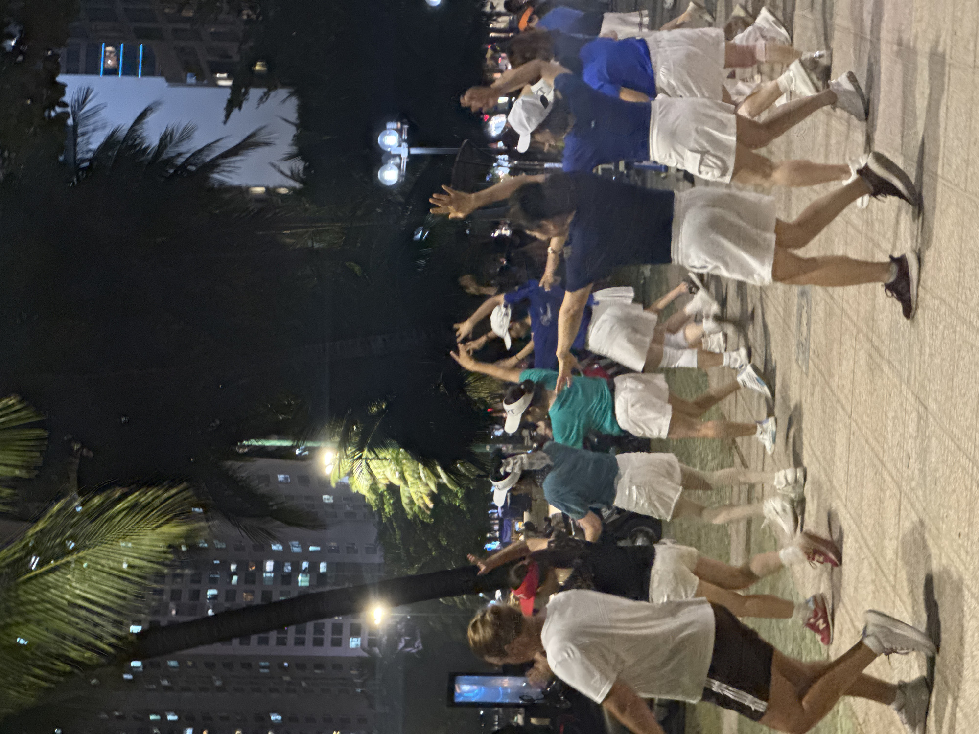 The image shows a row of people on a tiled pavement at night, with buildings and palm trees in the background. The people are wearing shorts and various colored t-shirts, some with white hats. They appear to be stretching or exercising, with arms raised and legs apart. Street lights illuminate the scene. The location is a city setting, judging by the buildings and the presence of street lights and pavements.