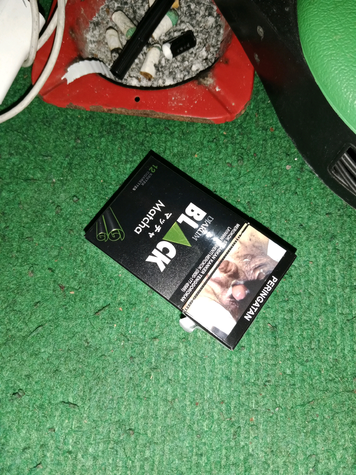 The image shows a close-up, angled shot of a black cigarette pack with the brand name "BLACK" and "Matcha" printed on it, lying on a green carpeted surface. The cigarette pack is partially open, revealing a warning label with a graphic image of a person's face, typical of tobacco product warnings in Indonesia. To the top left of the pack, a red ashtray filled with cigarette butts and ash is visible, along with a white electrical cord. The overall scene appears to be an indoor setting, possibly a residential area, with no specific landmarks indicating the city of Ringinrejo, Indonesia.