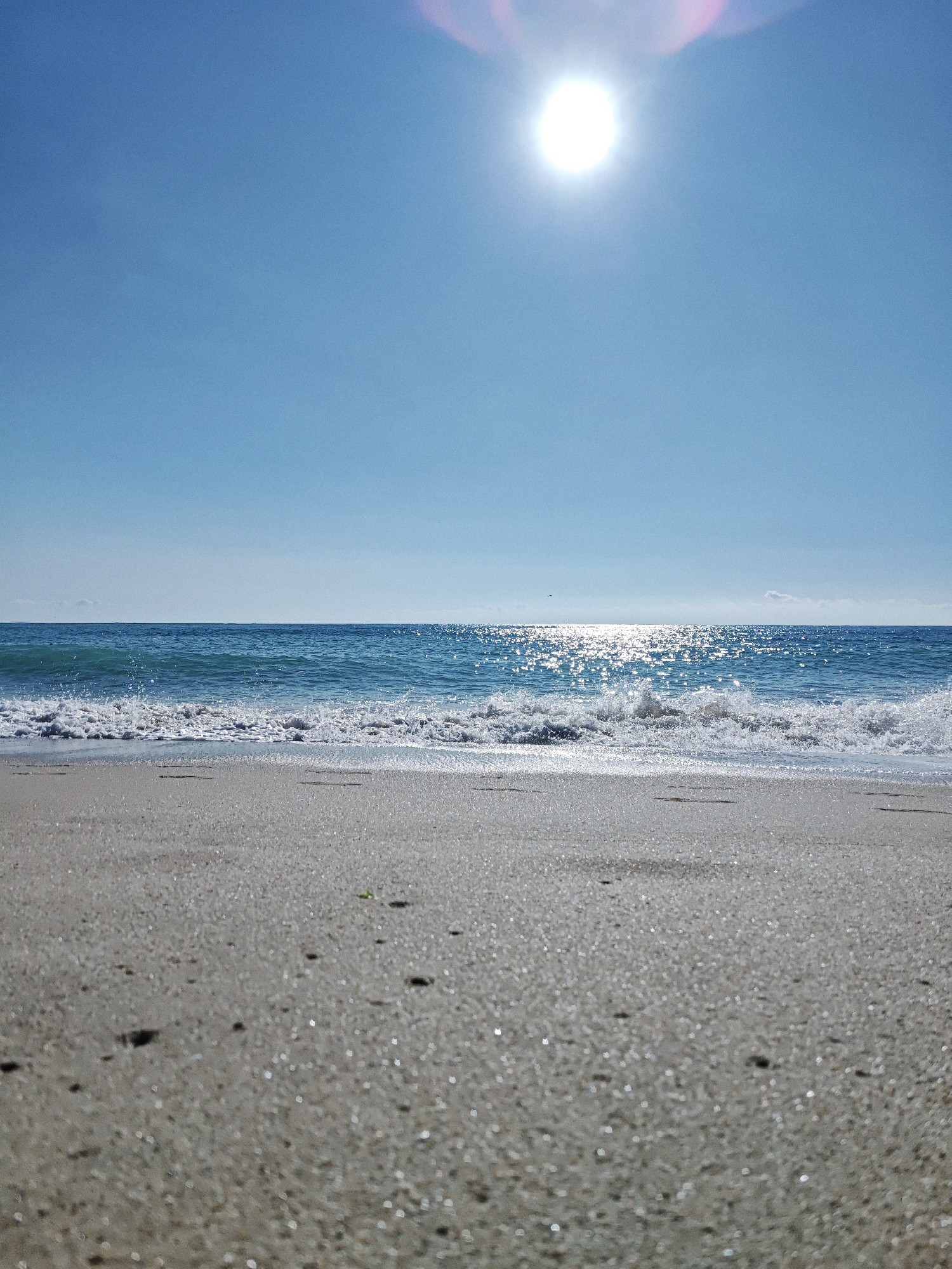A low-angle, vertical shot captures a sun-drenched beach scene. The foreground is dominated by a close-up view of wet sand, glistening with small, reflective particles, creating a sparkling effect. A few dark, irregular shapes, possibly small stones or debris, are scattered across the sand. In the mid-ground, a gentle wave breaks onto the shore, its white foam contrasting with the deep blue of the ocean. The water stretches out to a clear, unbroken horizon line under a bright, cloudless blue sky. High in the sky, a bright sun shines, its glare creating a soft, pinkish lens flare in the upper right quadrant of the image.