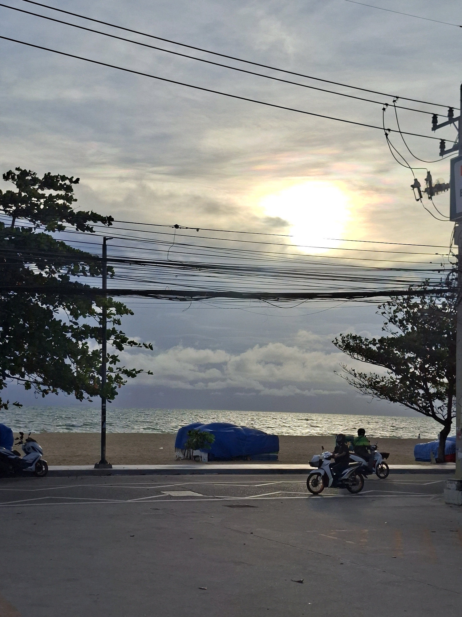 The image depicts a street scene in Pattaya City, Thailand, with the ocean visible in the background. The sky is overcast with the sun attempting to break through, creating a hazy, bright spot. Several utility wires are strung across the upper portion of the frame, supported by poles. On the left side of the image, a tree with green leaves is visible.
The scene includes a sandy beach adjacent to the water, with two blue tarpaulins covering objects on the sand. A dark-colored scooter is parked on the side of the road in the foreground on the left. Further down the road, two individuals are riding motorcycles. The road itself is paved, with white lane markings. The overall impression is of a coastal city street during the daytime, with a somewhat diffused light.