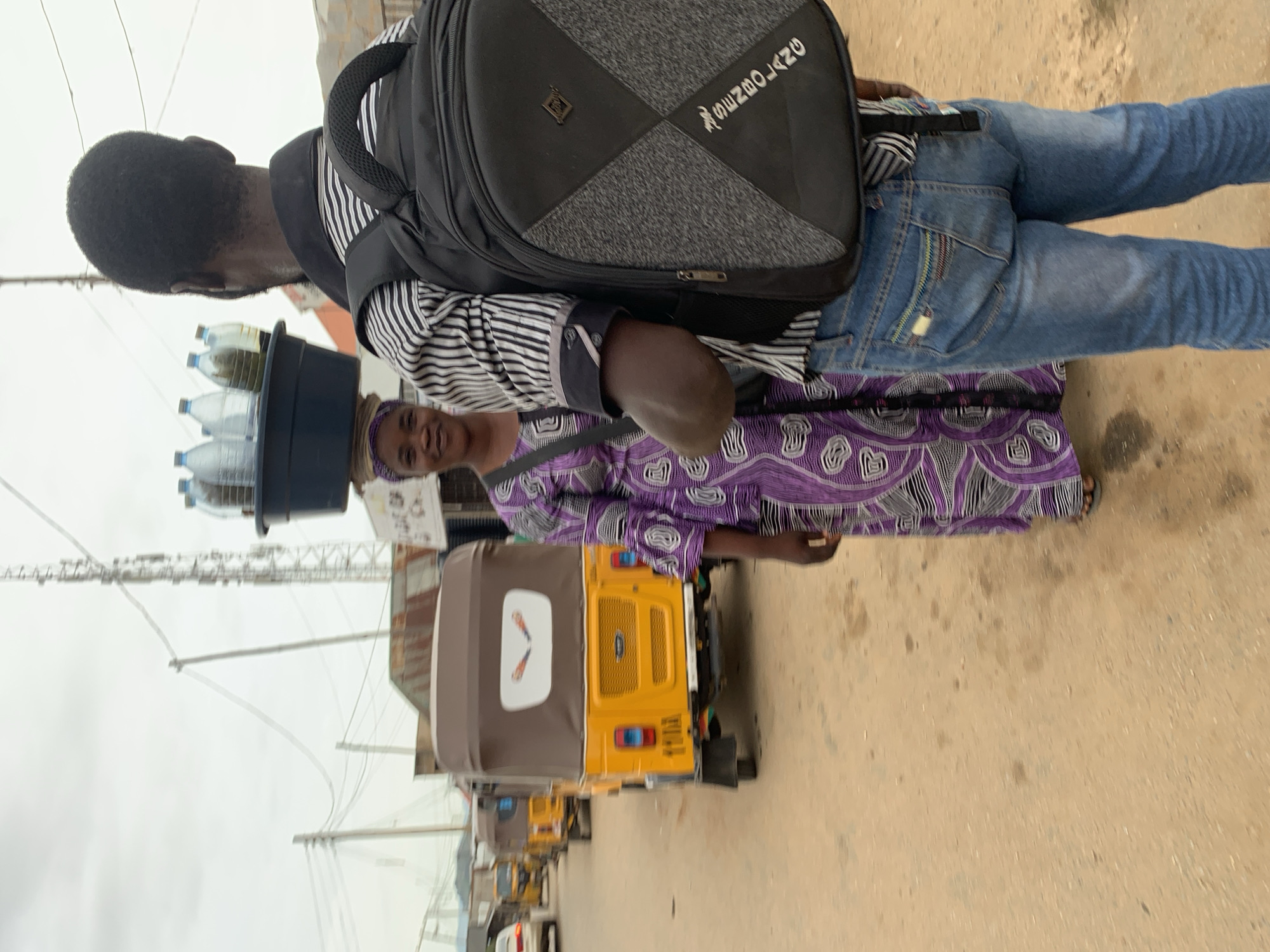 In Abuja, Nigeria, a woman, facing forward with a faint smile, balances a dark basin filled with clear and greenish liquid-filled bottles on her head. She wears a purple and white patterned traditional African dress and a dark headscarf. She stands in a dusty outdoor environment, with a man partially obscuring her from view. The man, seen from behind, wears a black and white striped shirt, blue jeans, and a black backpack with grey patterned sections. In the background, several yellow three-wheeled vehicles, commonly known as 'Keke Napep' or 'Maruwa', are parked on the side of a dirt road. The sky is overcast, and utility poles and wires are visible, suggesting an urban or semi-urban setting within Abuja.