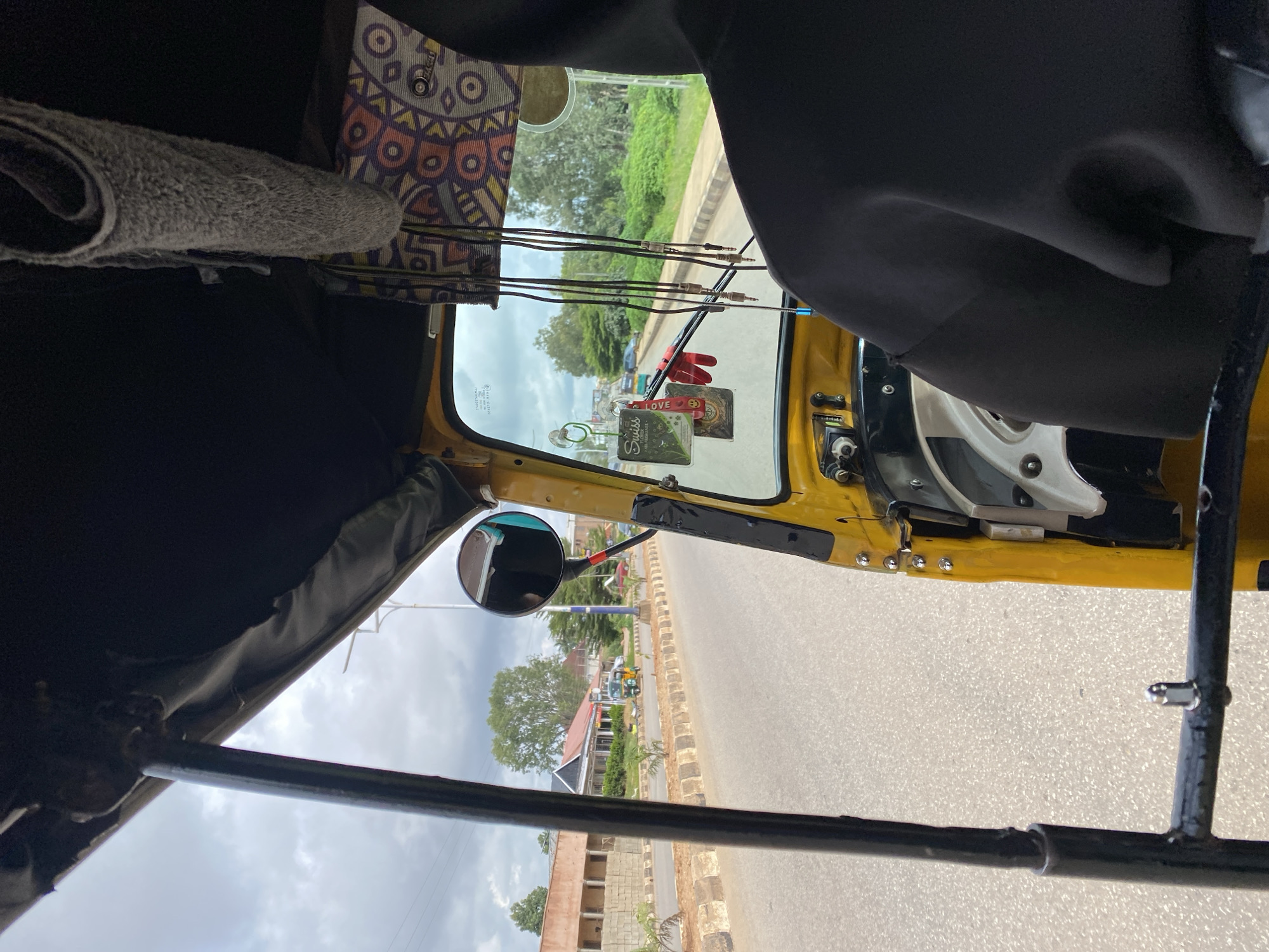 The image shows a view from inside a yellow auto-rickshaw, likely in Barakin Lerawi, Nigeria. The perspective is from the passenger seat, looking forward through the windshield. A person wearing a black hooded garment is seated in the foreground, facing forward.
The auto-rickshaw's interior features a black roof lining with a grey towel hanging down. Decorative fabric with a colorful geometric pattern is also visible above the windshield. Various cables and audio jacks hang from the ceiling. A side mirror is mounted on the right side of the windshield.
Outside the vehicle, a paved road is visible, bordered by a concrete curb and a sidewalk. Buildings with light-colored brick and red roofs are situated along the road. Green trees and vegetation are present in the background, under a cloudy sky. Other vehicles, including a green auto-rickshaw and cars, are on the road. The overall scene depicts a moment of travel within a Nigerian town or city.