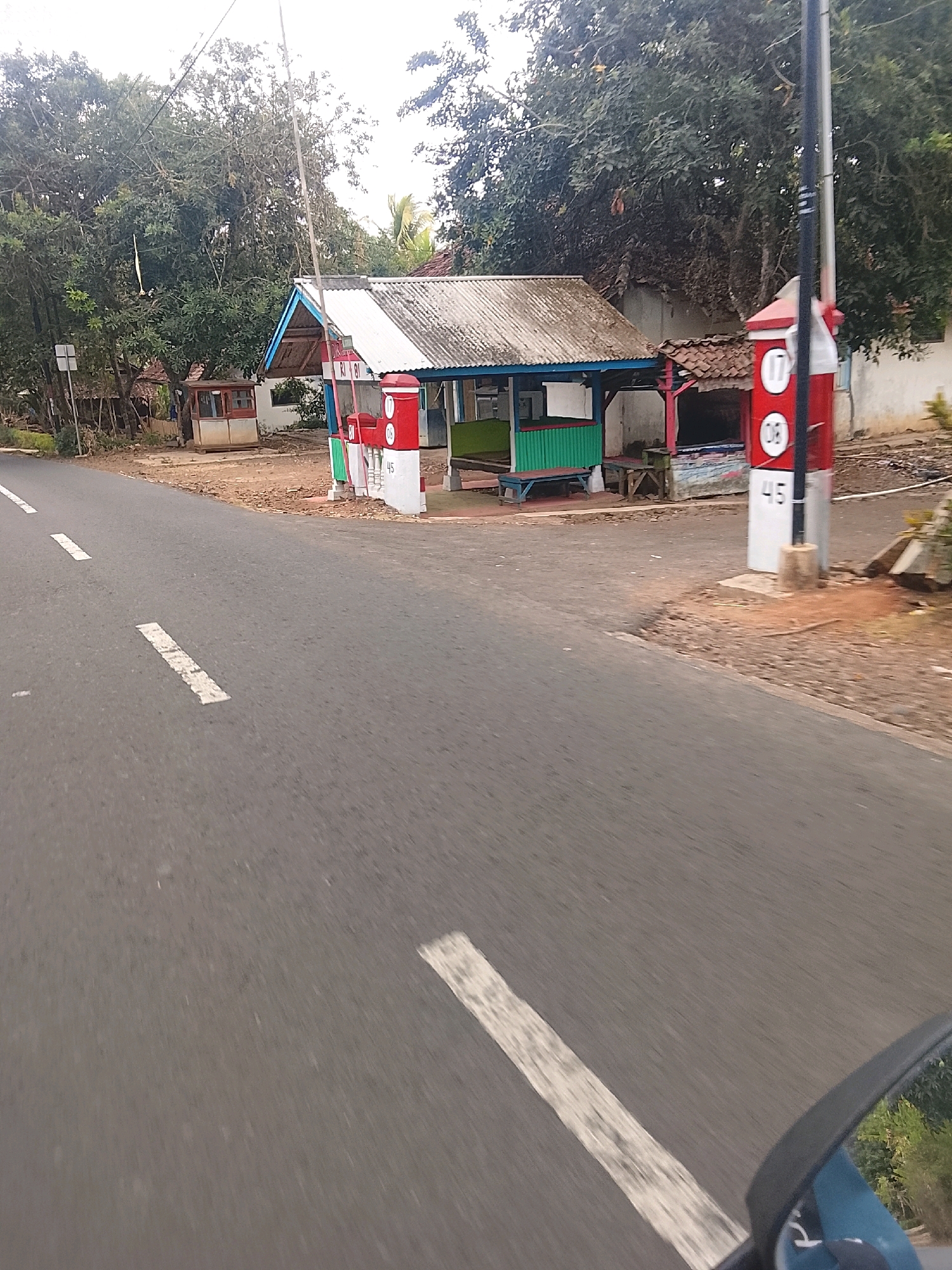 The image depicts a roadside scene in Banjar, Indonesia. In the foreground, a paved road runs diagonally across the frame, marked with white lines. The environment includes a small, open-air structure with a corrugated metal roof, likely a roadside stall or kiosk, featuring green and white corrugated metal siding. Adjacent to it is a red and white marker post with circular elements containing the numbers "17" and "08", and a lower section with the number "45". Trees and foliage surround the structures. The overall impression is of a rural or semi-urban setting. There are no people visible in the image.