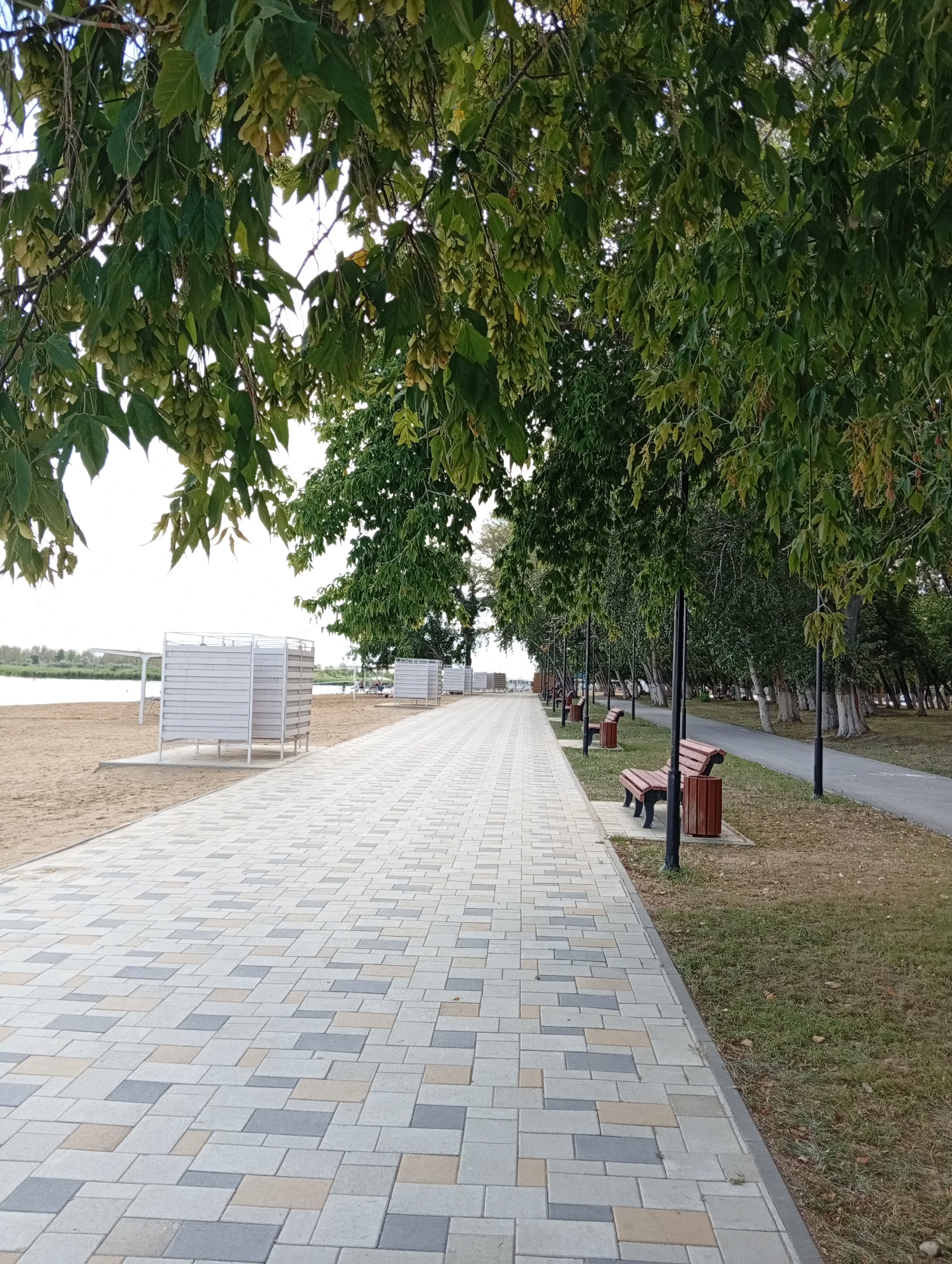 The image depicts a paved walkway on the left, running alongside a sandy beach with several white, slatted changing rooms. On the right of the walkway, there is a strip of grass with a secondary paved path running parallel to it. This secondary path is lined with lamp posts and wooden benches. A dense row of trees with green foliage borders the grassy area, creating a shaded environment. In the distance, a body of water is visible beyond the beach. The scene appears to be a public recreational area, likely a beachside promenade. The location is identified as Michurinsk Rural District, Kazakhstan. There are no people clearly visible in the image.